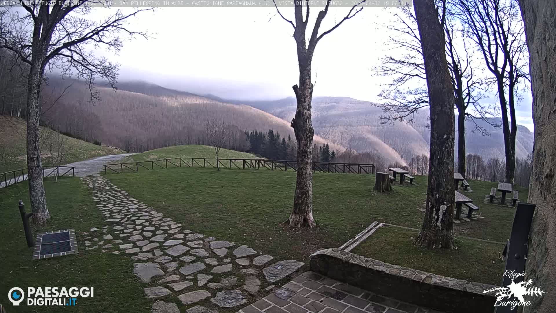 A stone pathway leads to picnic tables situated on a grassy area overlooking a mountain range partially obscured by fog.