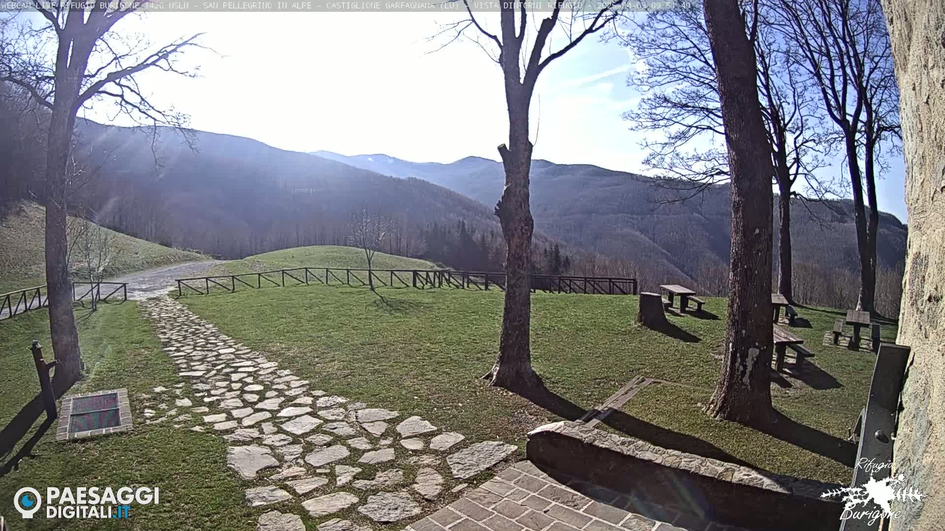 A stone path leads to several picnic tables situated on a grassy area overlooking a mountain range under a sunny sky.