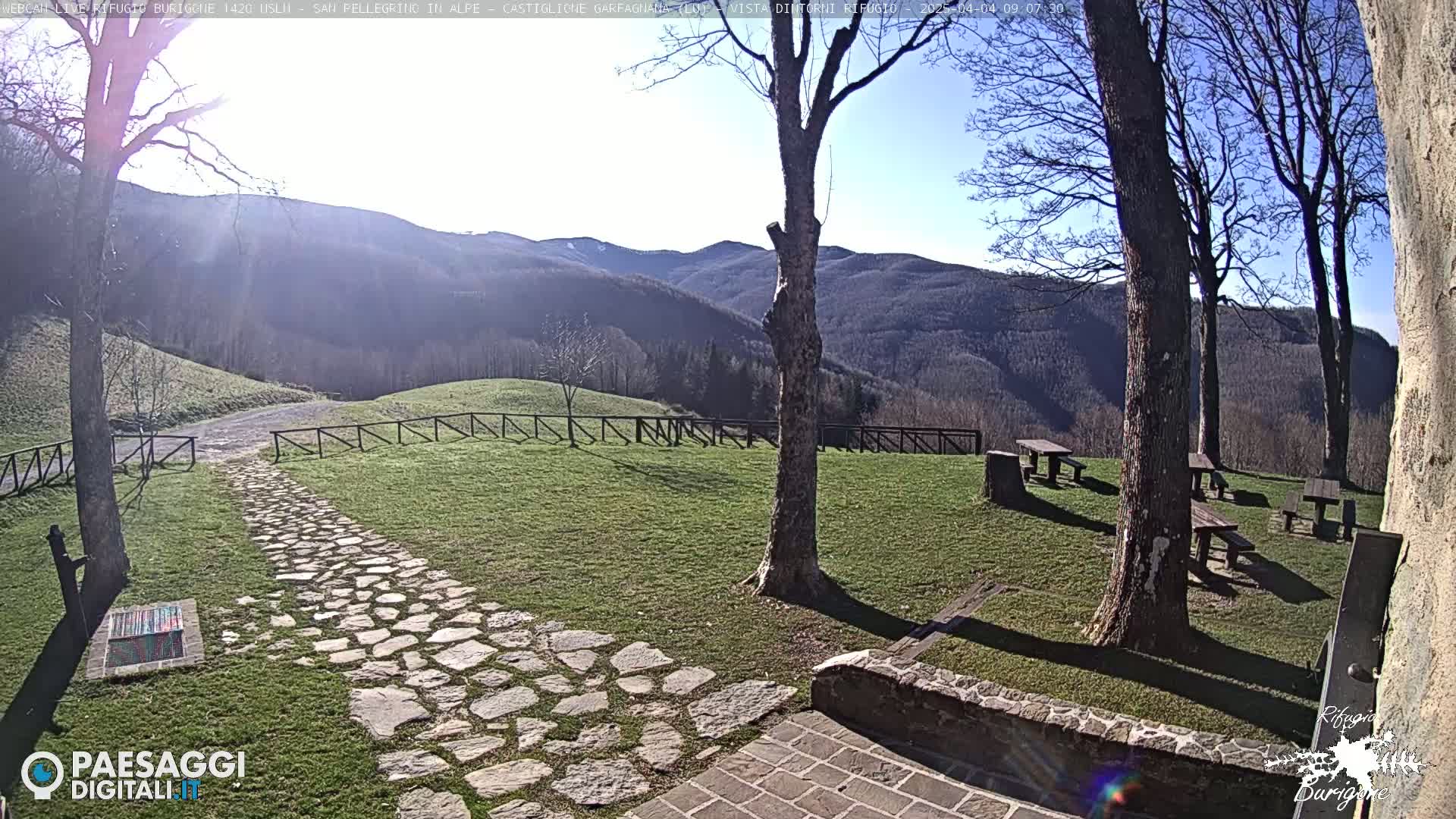 A stone path leads to picnic tables in a grassy area overlooking a mountain range on a sunny day.