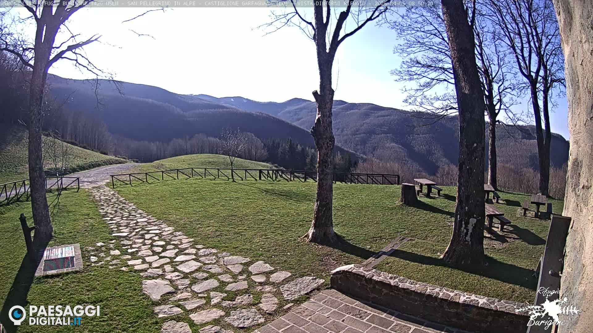 A stone path leads to picnic tables situated on a grassy area overlooking a mountain range under a clear, sunny sky.
