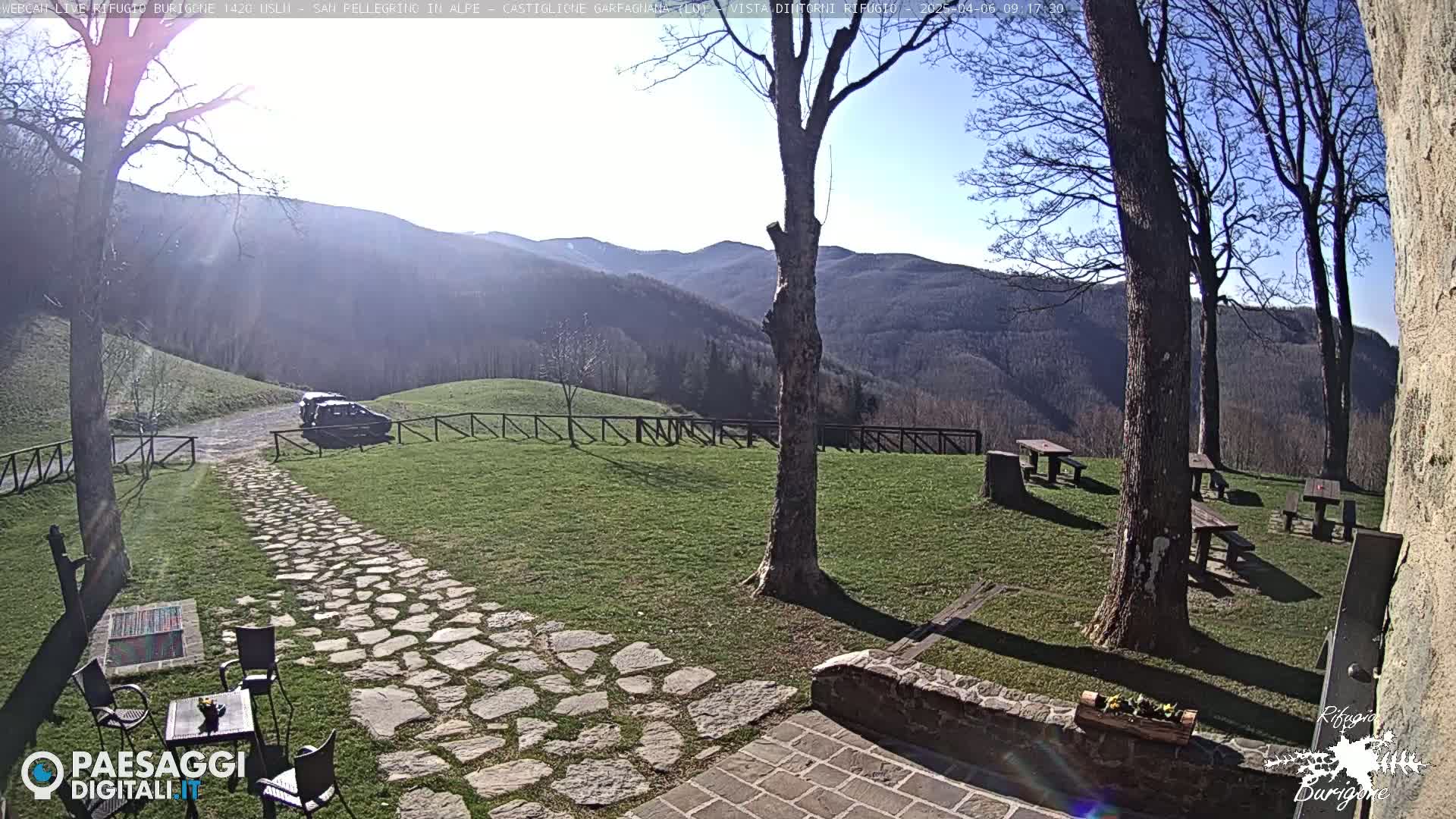 A stone path leads to a parked car in a grassy area with several picnic tables, trees, and a mountain view under sunny skies.