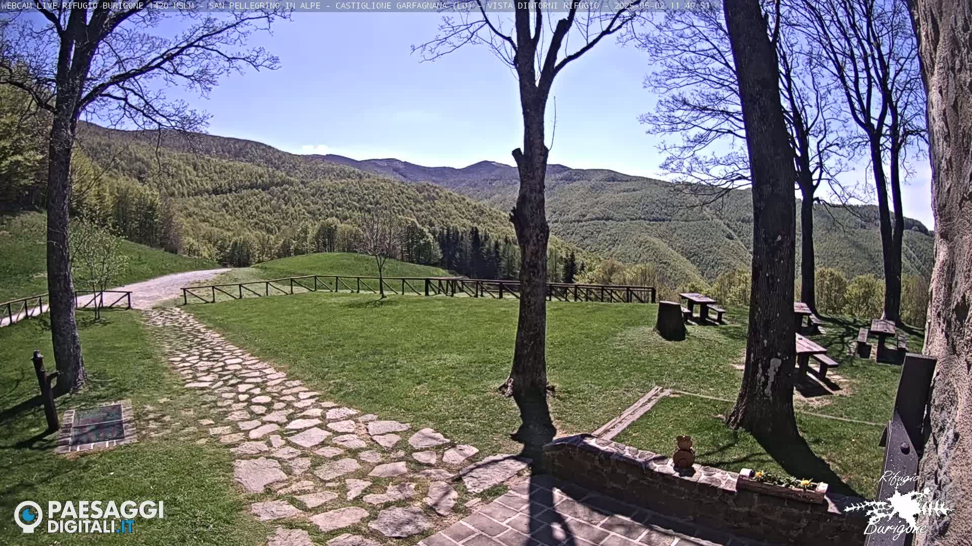 A stone path leads to a grassy area with picnic tables, surrounded by trees and overlooking a mountain range under a clear blue sky.