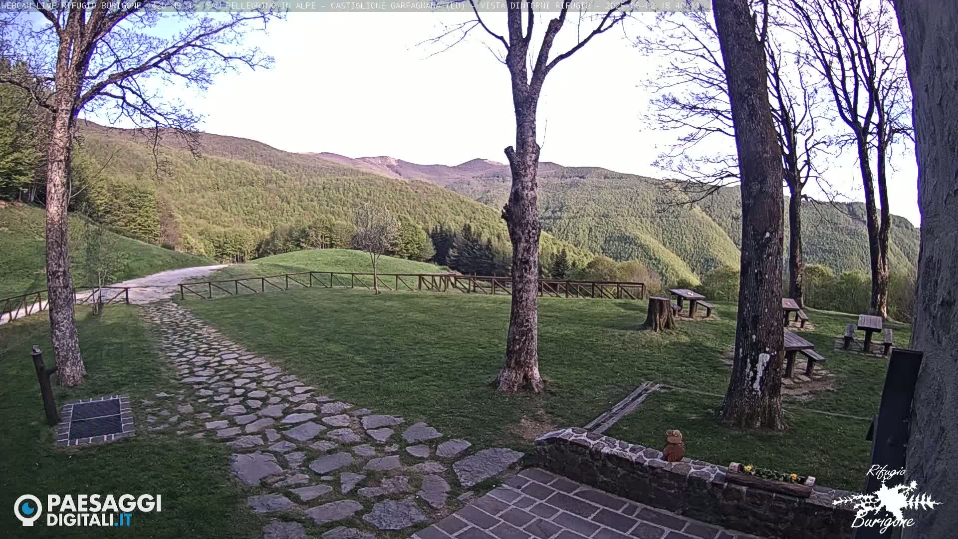 A stone path leads to picnic tables situated on a grassy area, overlooking a verdant hillside under a mostly clear sky.