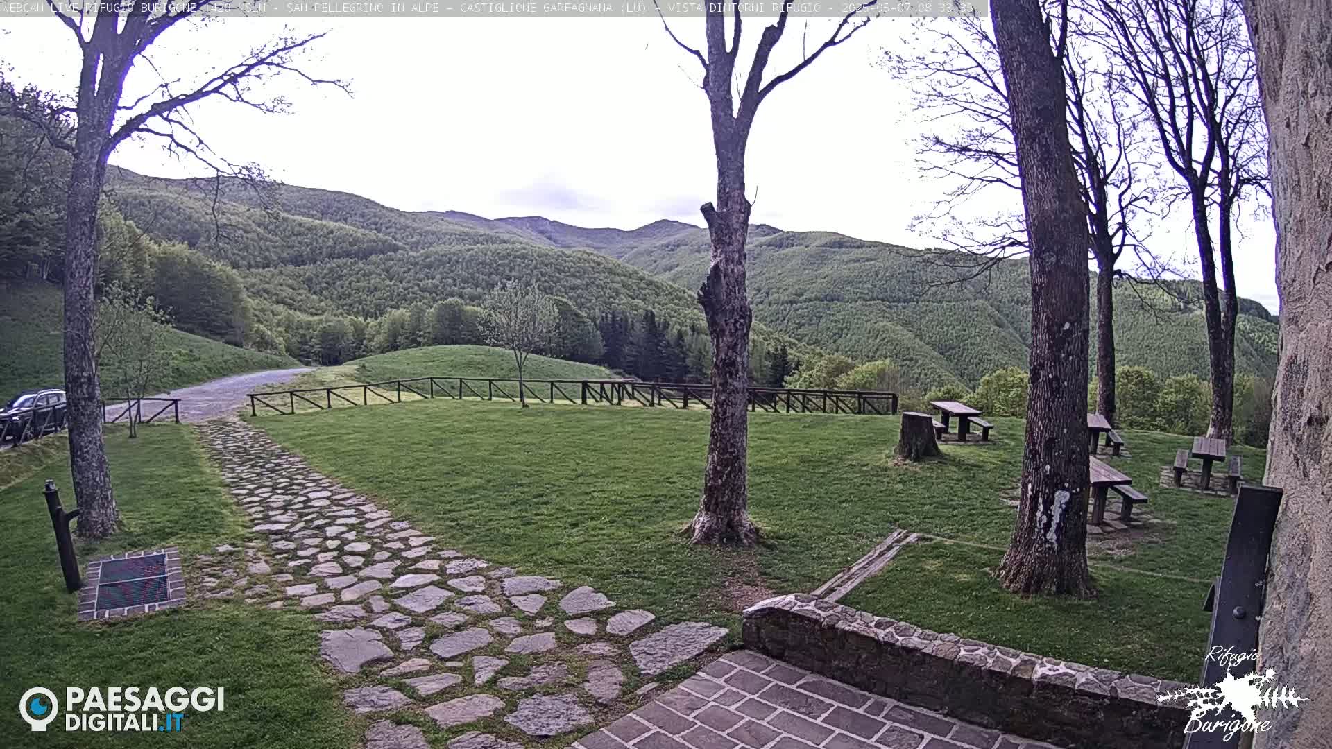 A stone path leads to several picnic tables situated on a grassy area overlooking a verdant hillside under a partly cloudy sky.