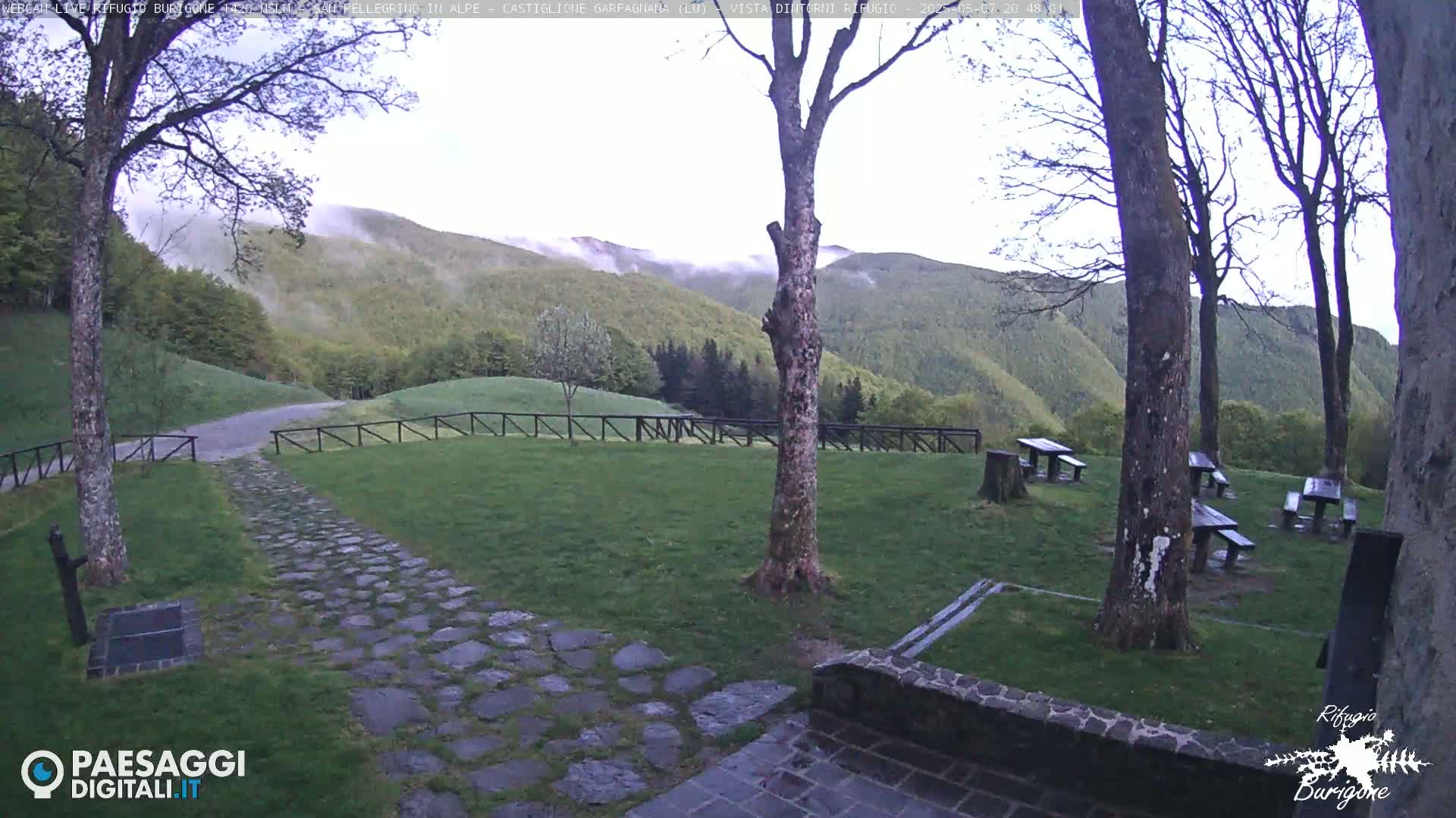 A stone path leads to a grassy area with picnic tables, surrounded by trees and overlooking a misty, verdant mountain range.