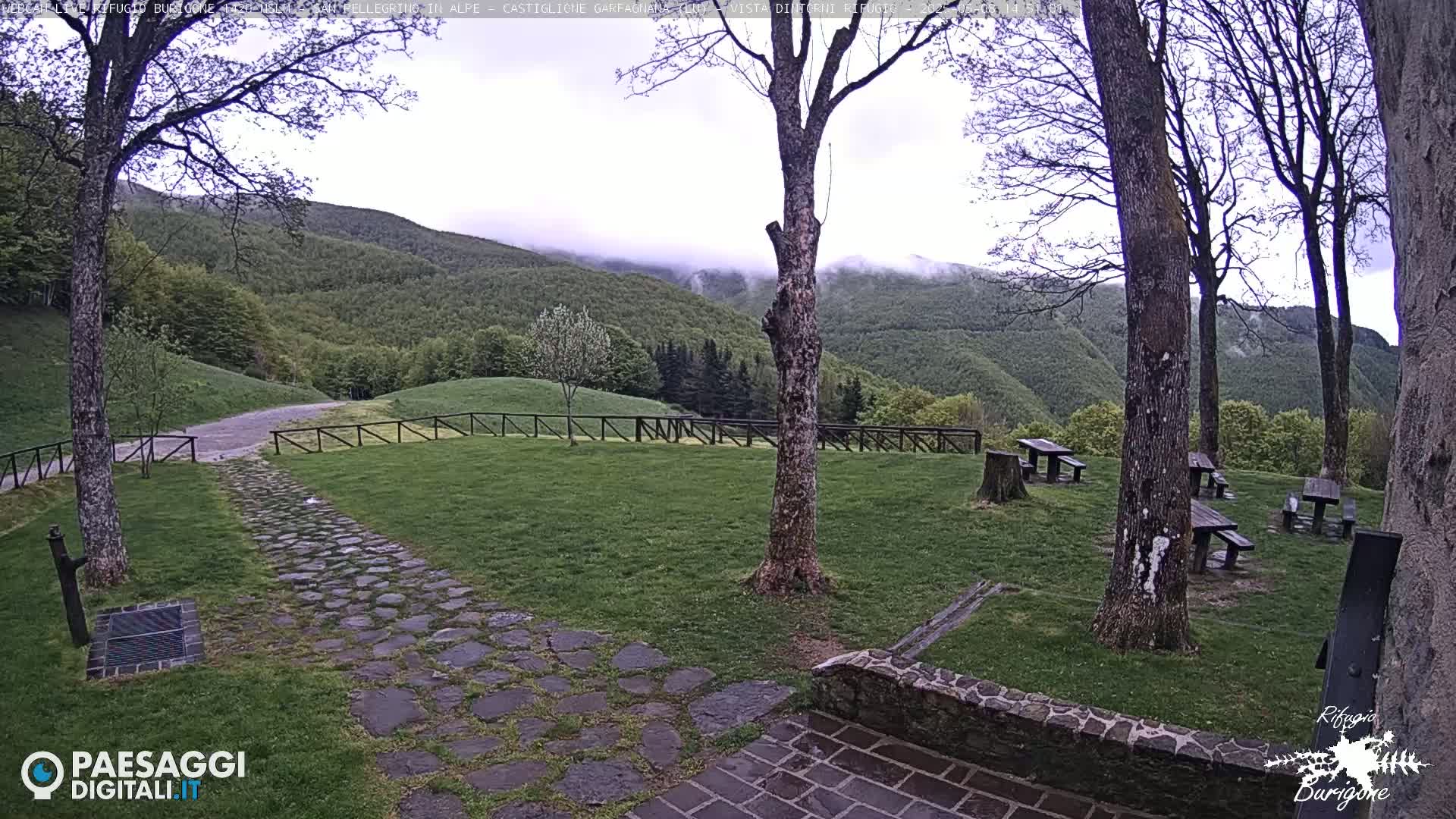 A stone path leads to picnic tables in a grassy area with a wooden fence overlooking a misty, forested mountain range.
