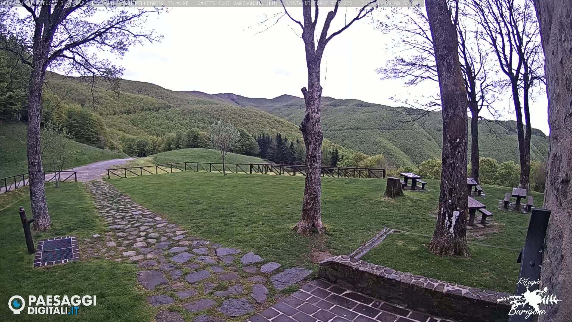 A stone path leads to picnic tables situated on a grassy area overlooking a lush green mountainside under a partly cloudy sky.