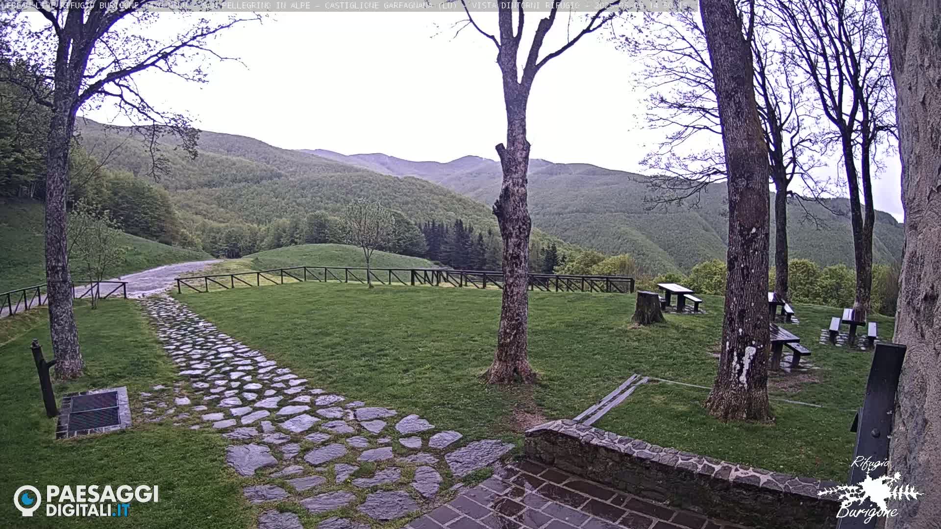 A stone path leads to picnic tables situated on a grassy area overlooking a verdant mountain range under an overcast sky.