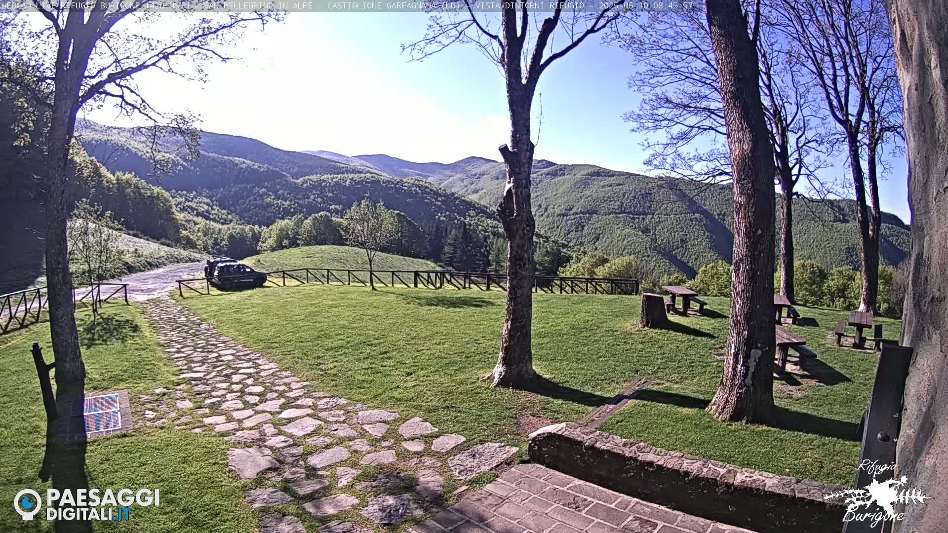 A stone path leads to a grassy area with picnic tables and a parked vehicle, all set against a backdrop of rolling green hills under a clear blue sky.