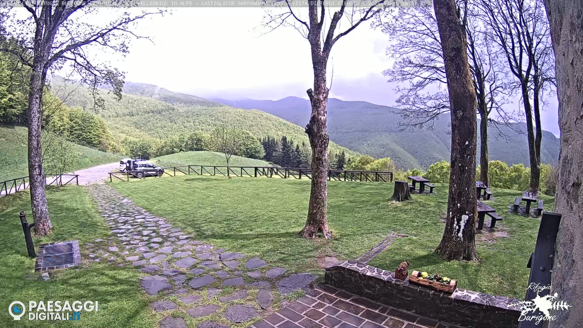 A stone path leads to a grassy area with picnic tables and a parked vehicle, overlooking a mountainous landscape under an overcast sky.