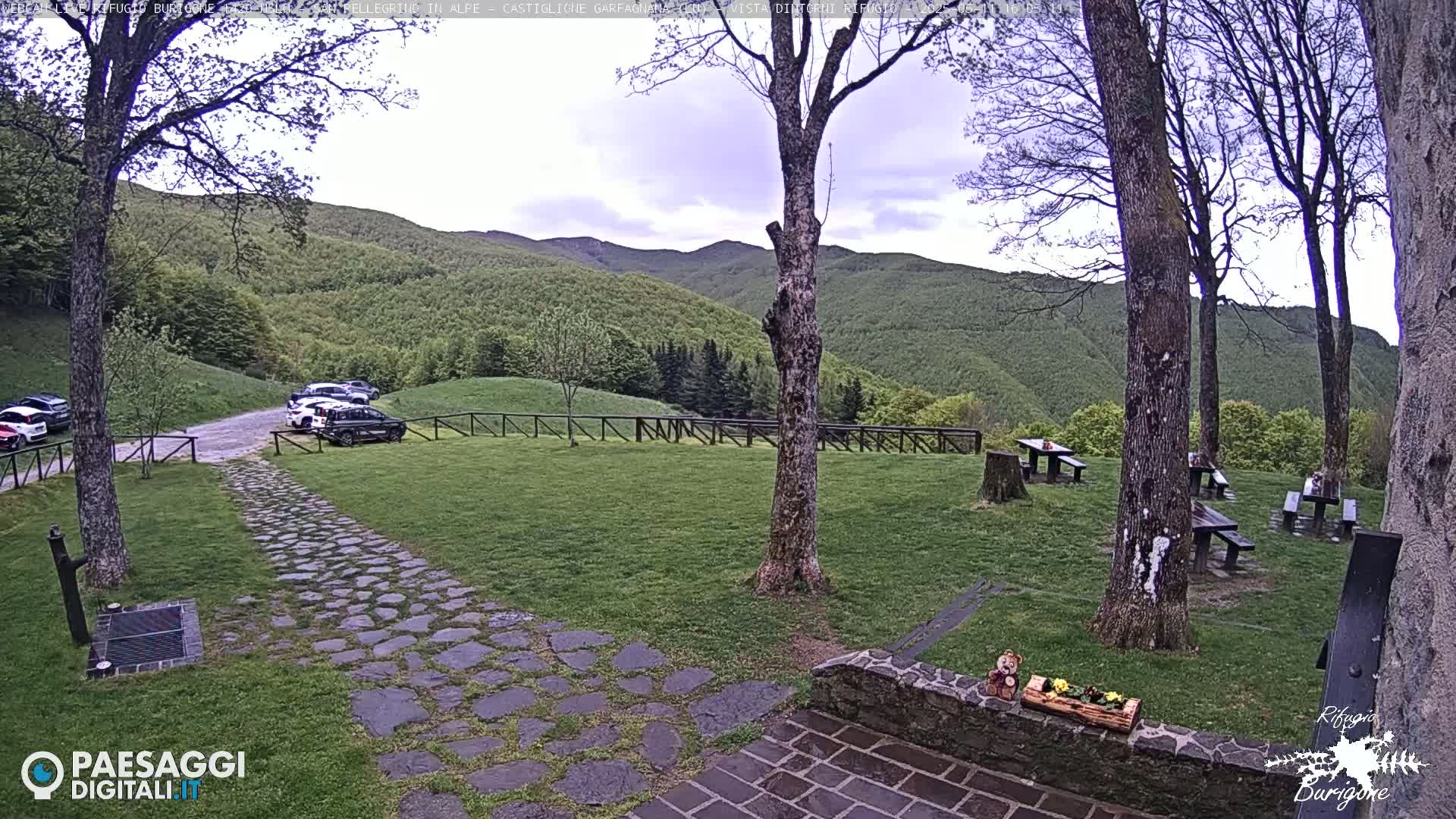 A stone path leads to a grassy area with picnic tables and a parking lot, all overlooking a verdant mountain range under a partly cloudy sky.