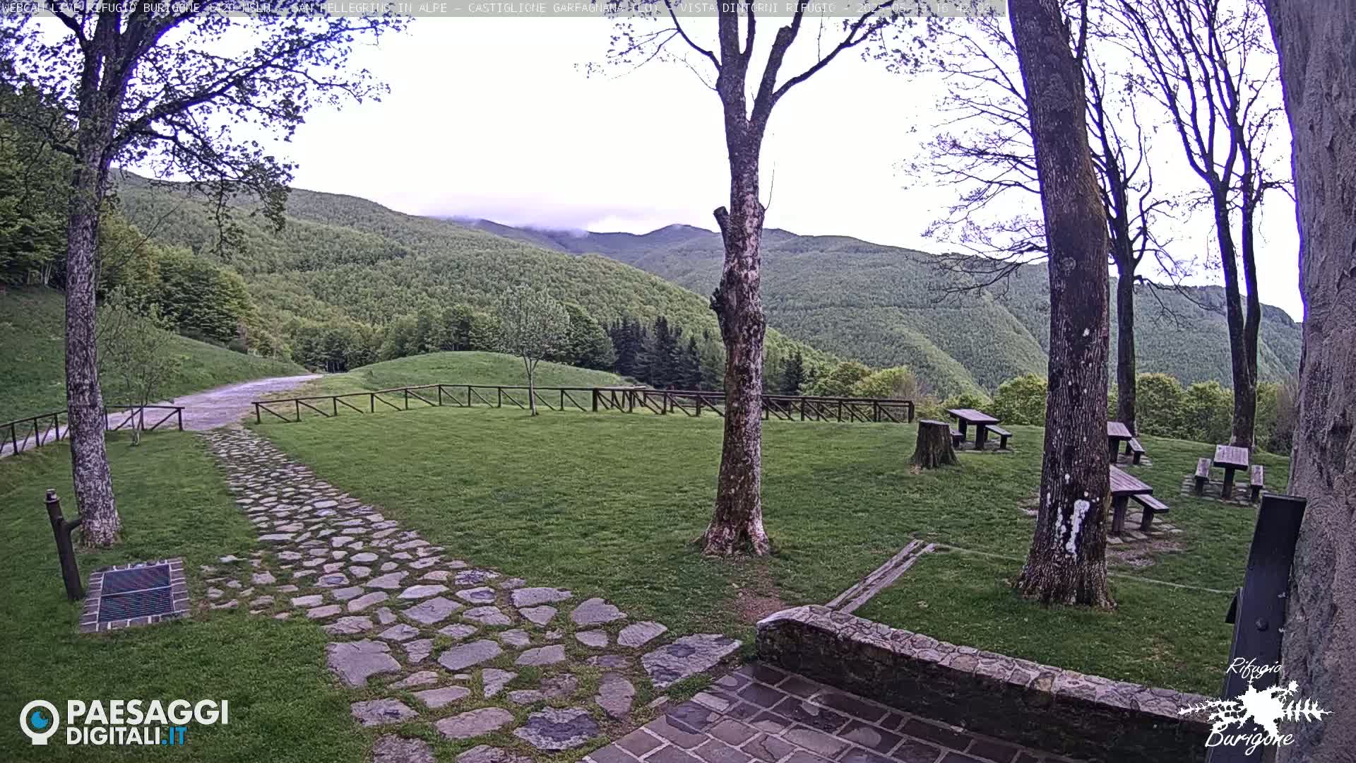 A stone path leads to several picnic tables situated on a grassy area, with a background of lush green hills and an overcast sky.