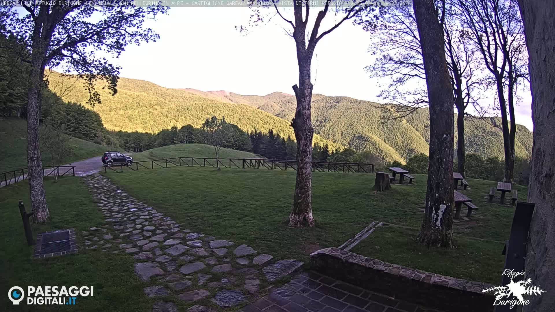 A stone path leads to a dark-colored SUV parked near a wooden fence, with several picnic tables scattered across a grassy area overlooking a mountain range under a partly clear sky.