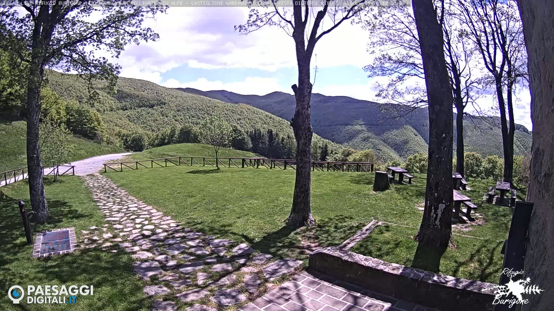 A stone path leads to a grassy area with picnic tables, surrounded by trees and hills under a partly cloudy sky.