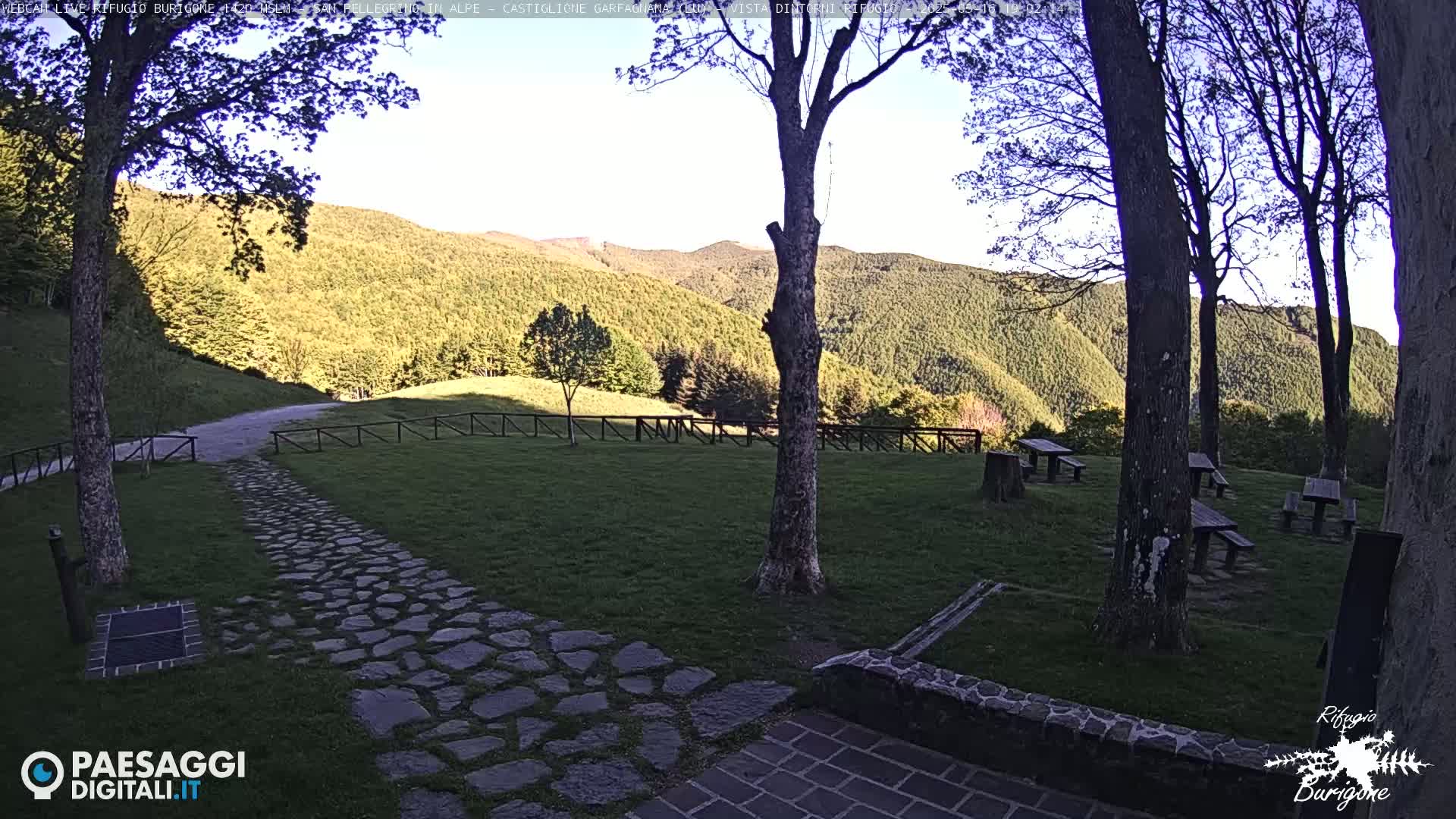 A stone path leads to several picnic tables situated on a grassy area overlooking a mountainside under a partly sunny sky.
