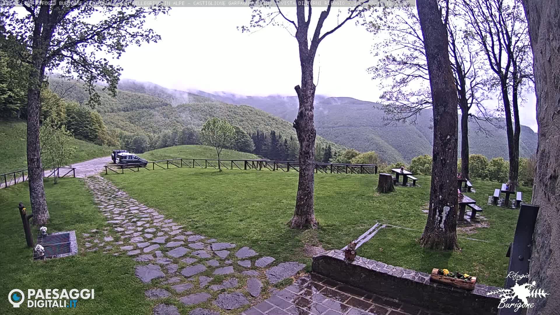 A stone pathway leads to a parking area with a car, beyond which is a grassy lawn with picnic tables, all set against a backdrop of misty, rolling green hills.