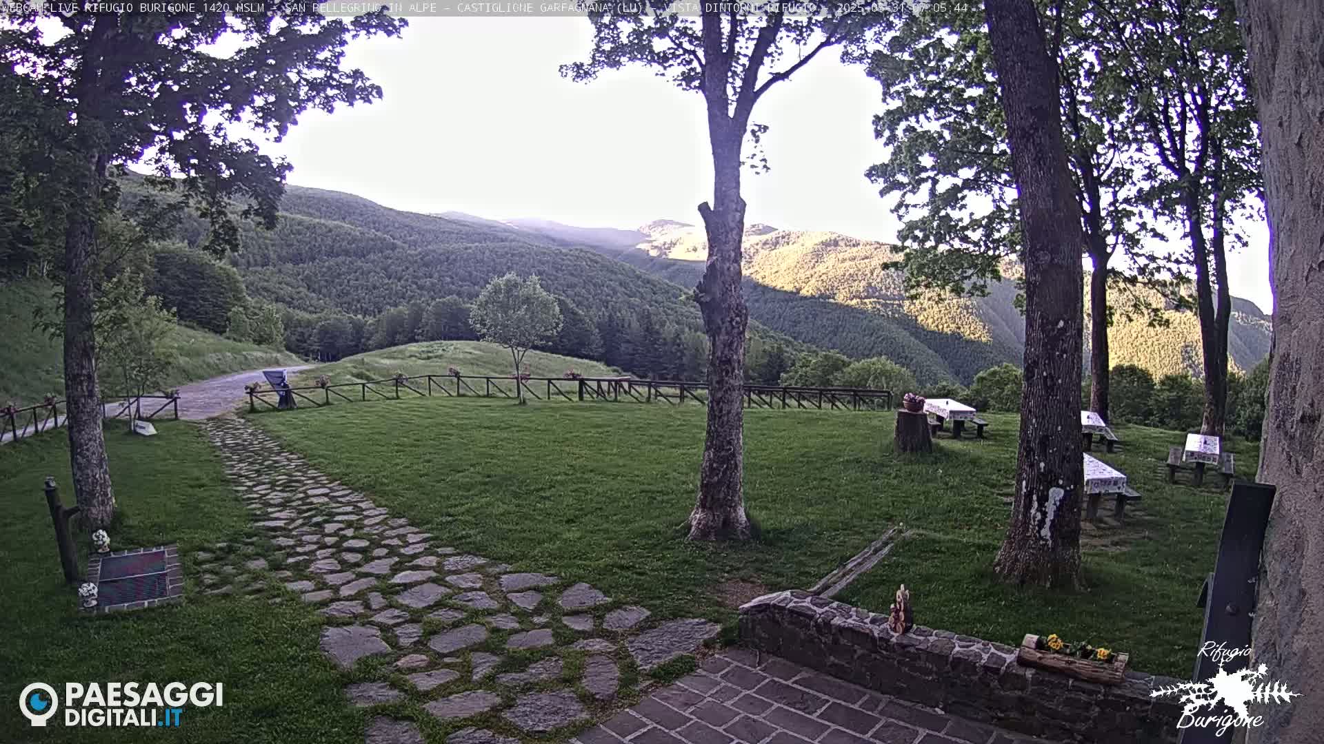A stone path leads to a grassy area with picnic tables under trees, overlooking a mountain range in the soft light of early morning.