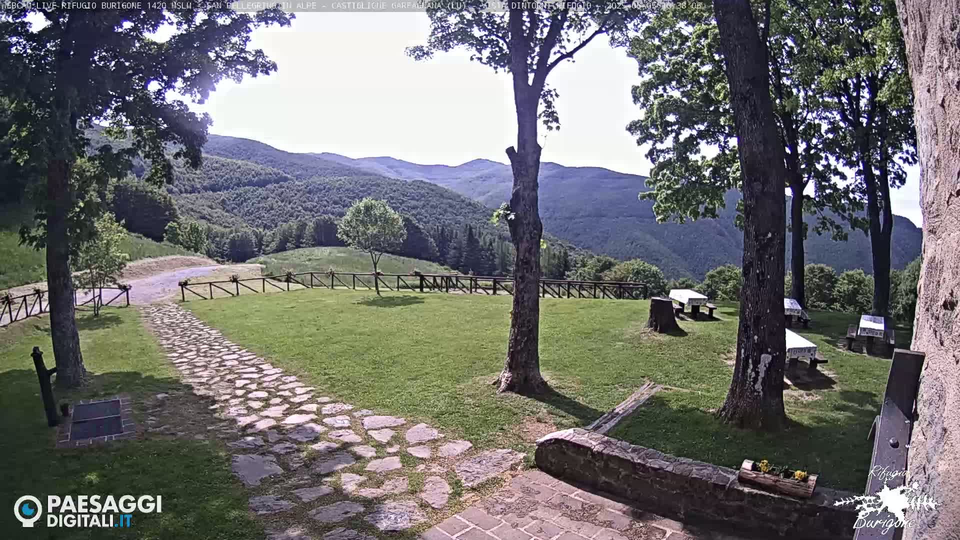 A stone path leads to a grassy area with picnic tables, trees, and a wooden fence overlooking a mountain range under a mostly sunny sky.