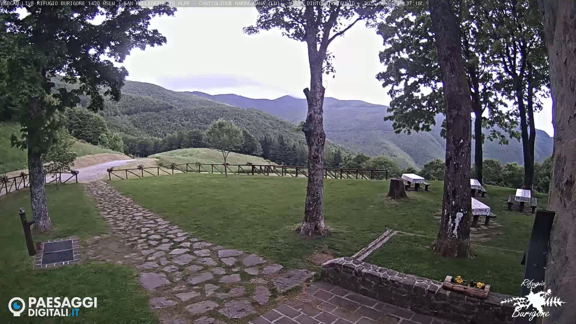 A stone path leads to a grassy area with several picnic tables under trees, overlooking a mountain range under an overcast sky.