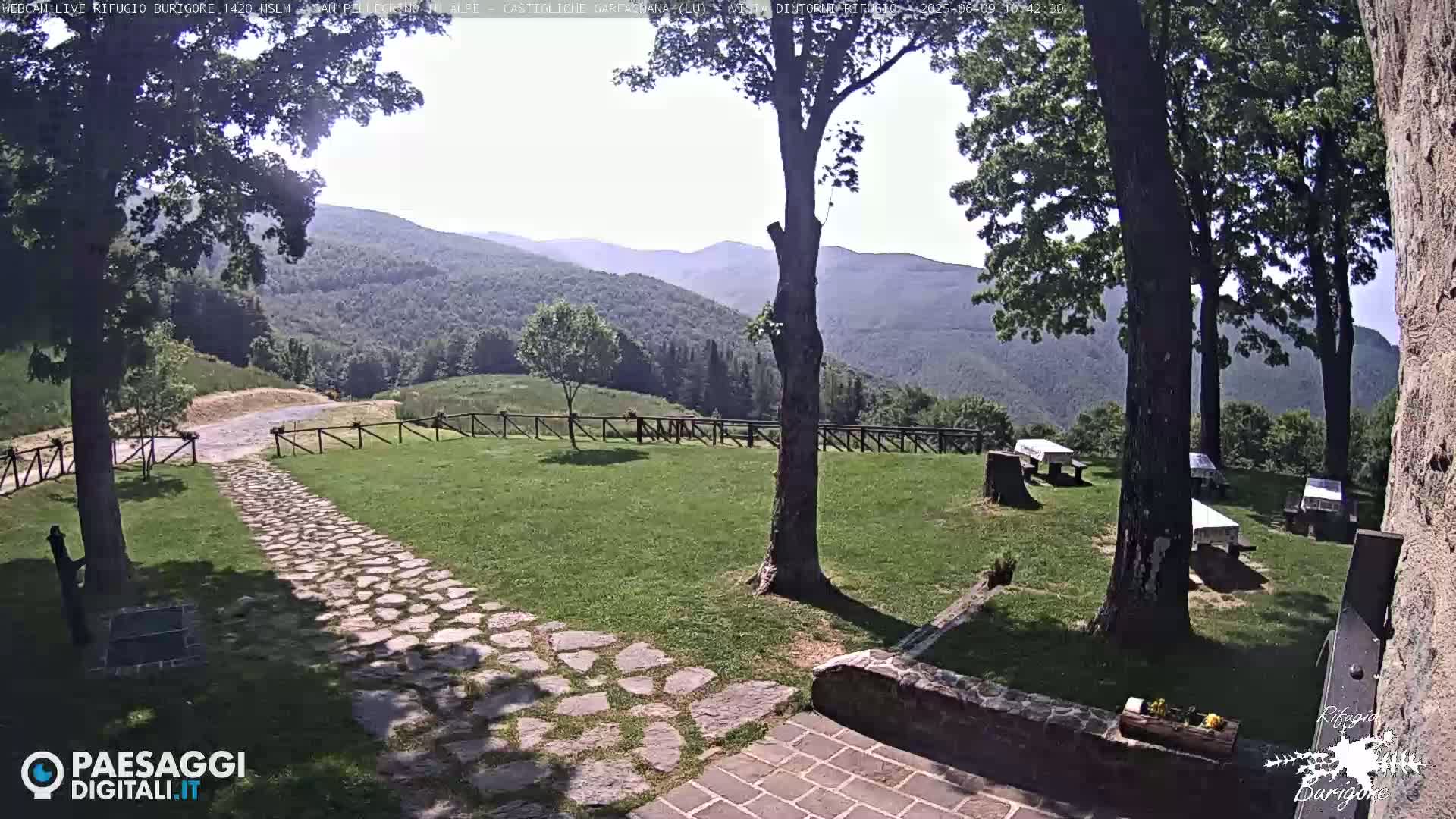 A stone path leads to a grassy area with picnic tables situated beneath trees, overlooking a mountainous landscape under a sunny sky.
