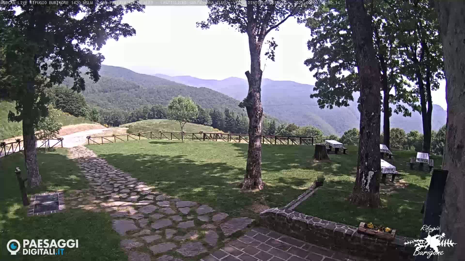 A stone pathway leads to a grassy area with picnic tables under trees, overlooking a mountain range on a sunny day.