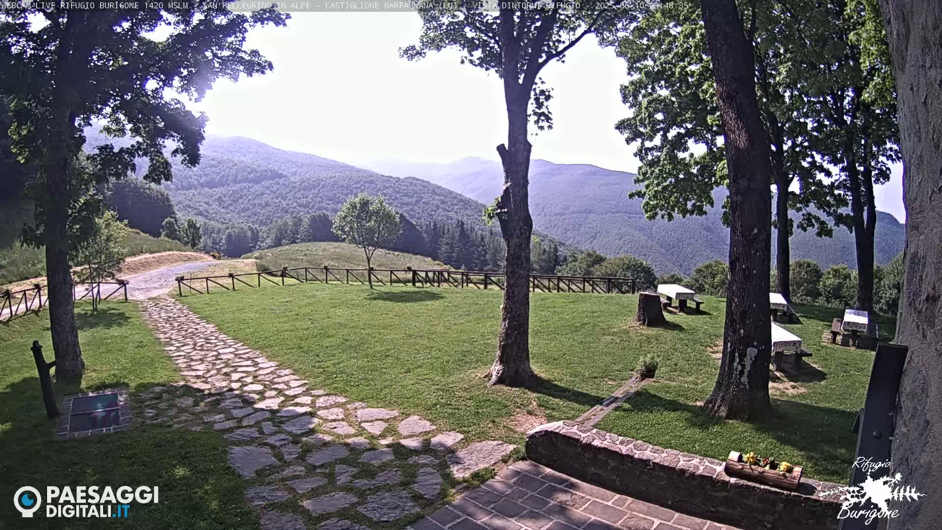 A stone path leads to a grassy area with picnic tables and trees, all overlooking a mountain range under a sunny sky.