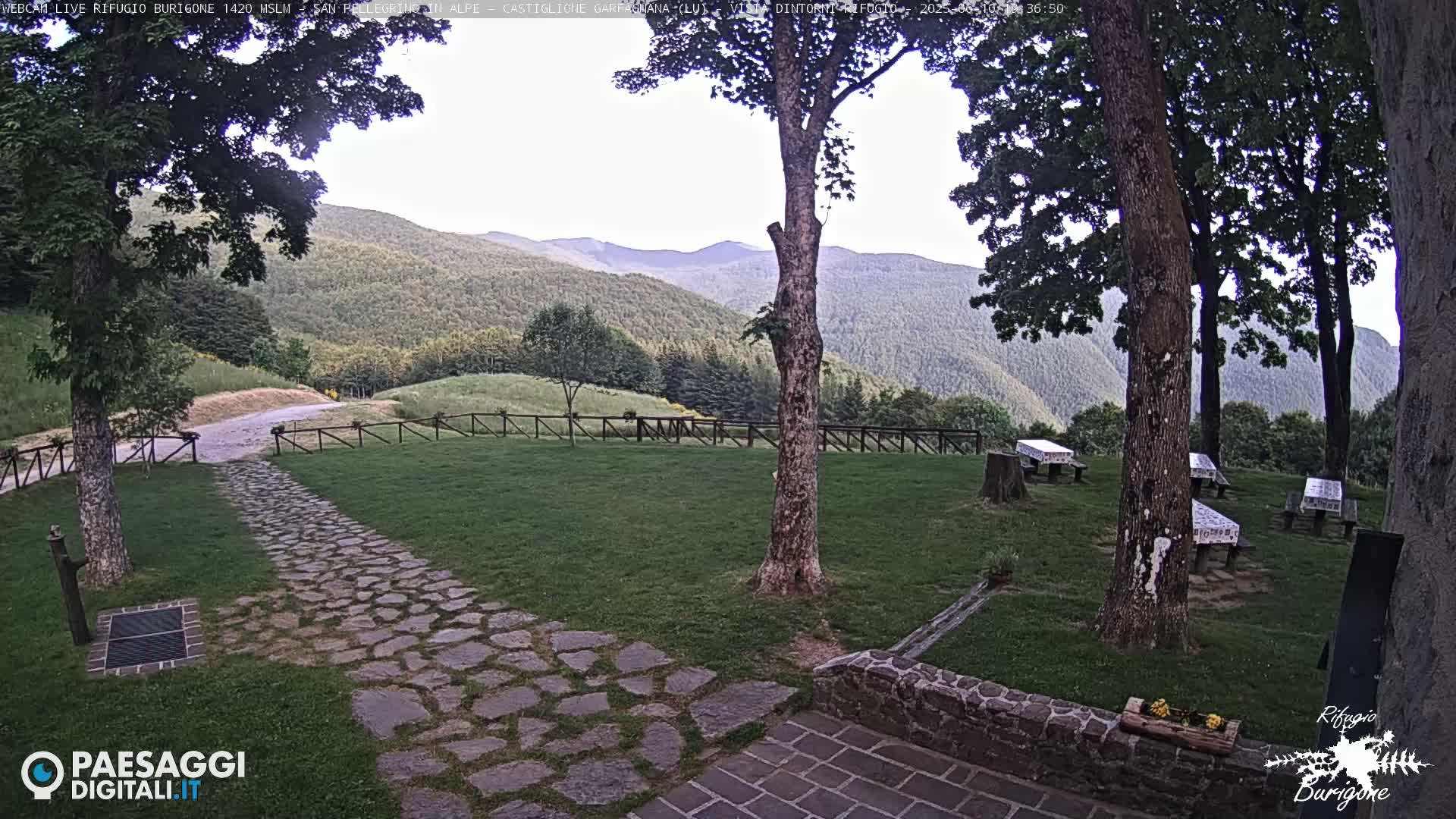 A stone pathway leads to picnic tables situated on a grassy area, surrounded by trees and overlooking a mountain range under a partly cloudy sky.