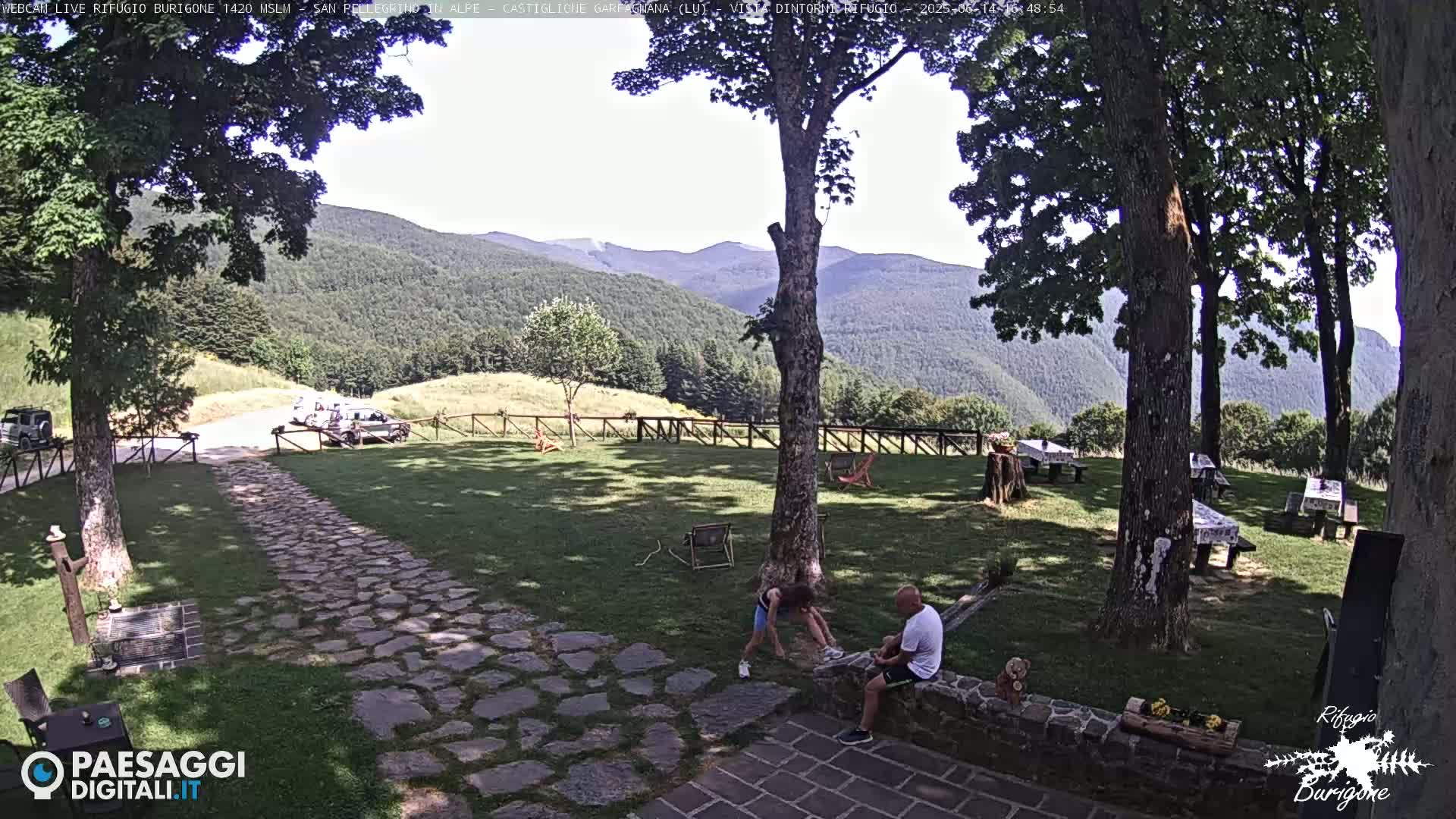 A grassy area with stone pathways, picnic tables, and several large trees overlooks a mountain range on a sunny day.