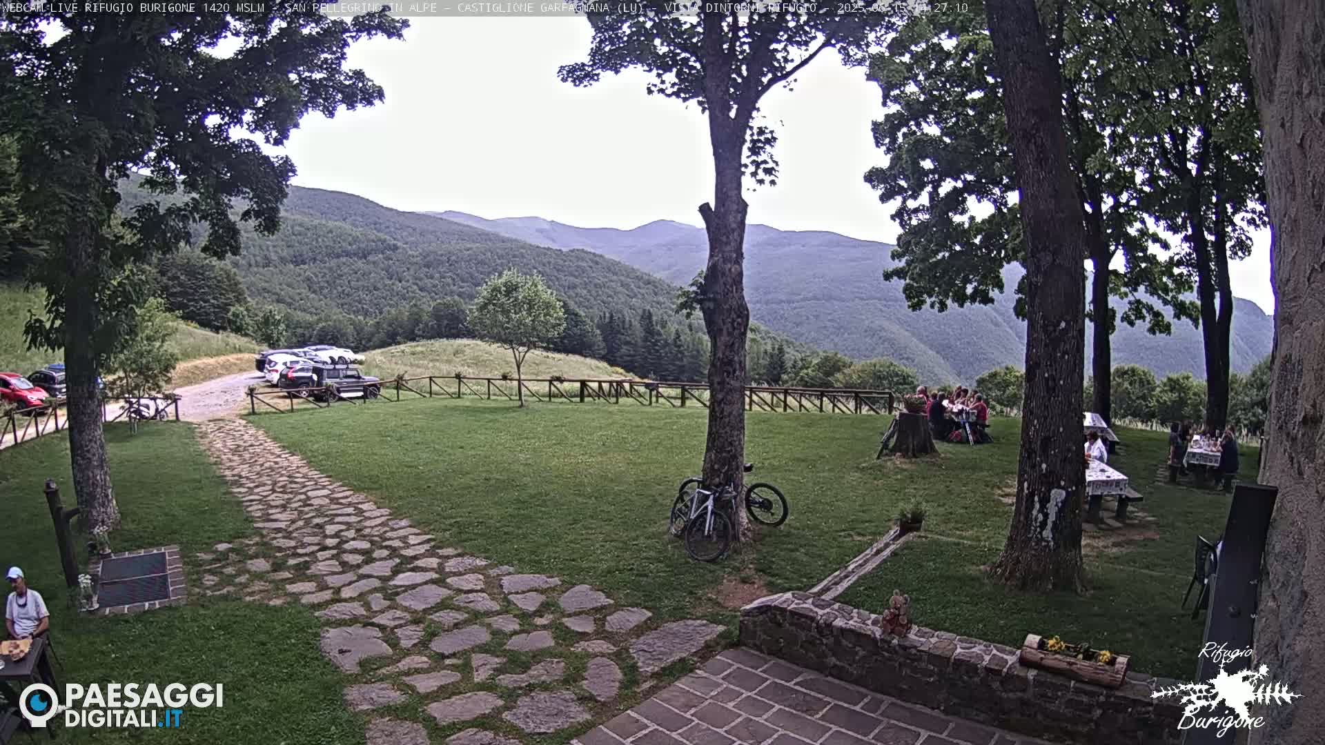 A stone path leads to a grassy area with several picnic tables surrounded by large trees, where people are seated, and cars parked in a lot in the background, all under an overcast sky with mountains visible in the distance.