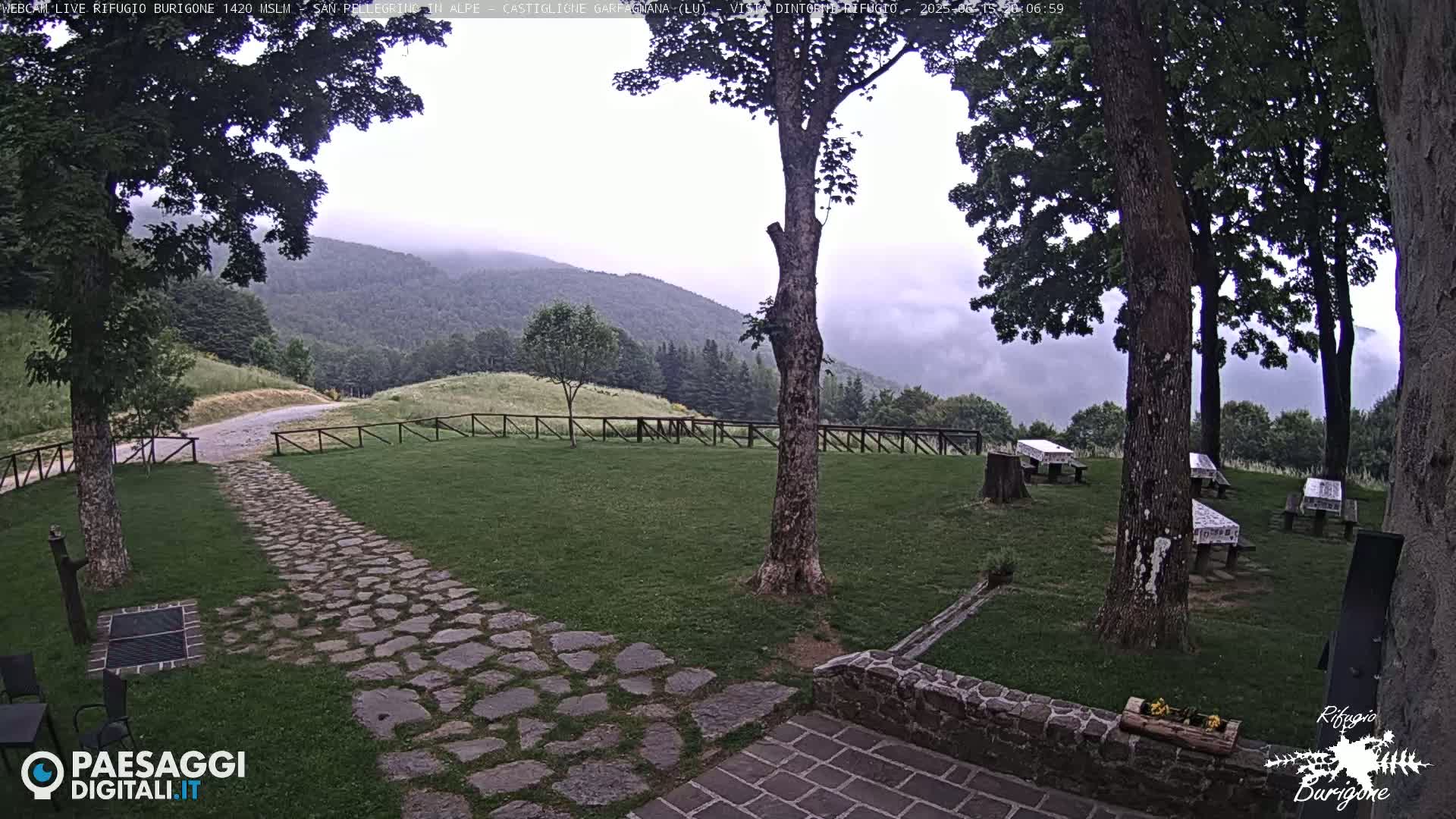 A stone path leads to a grassy area with picnic tables under trees, overlooking a misty mountain range.