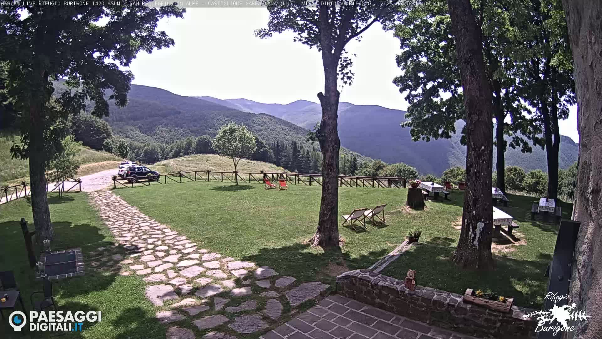 A stone path leads to a grassy area with picnic tables and lounge chairs, situated in front of a mountainous landscape under a mostly sunny sky.