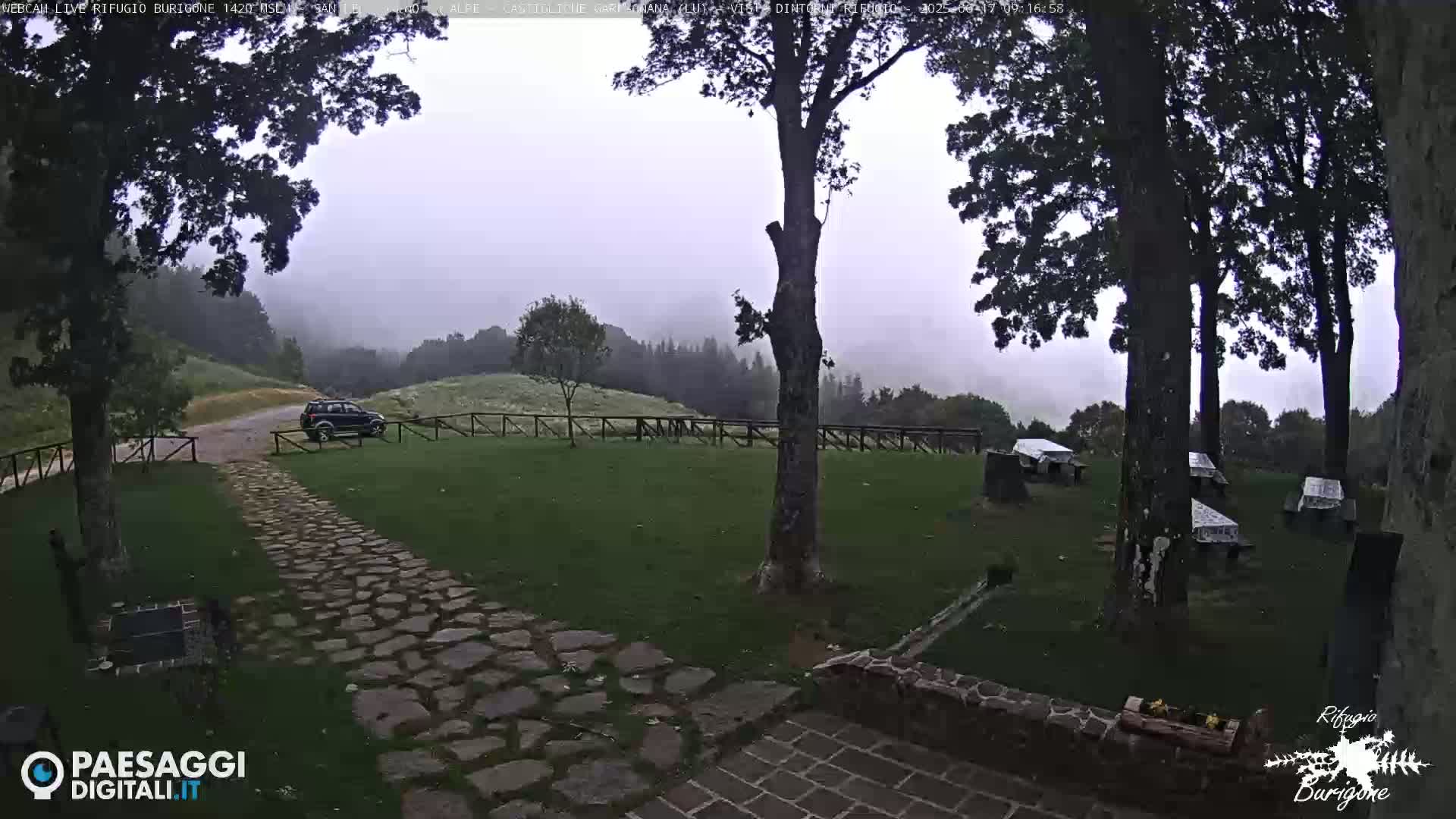 A stone path leads to a dark-colored SUV parked near a grassy area with picnic tables under a foggy sky in a mountainous location.