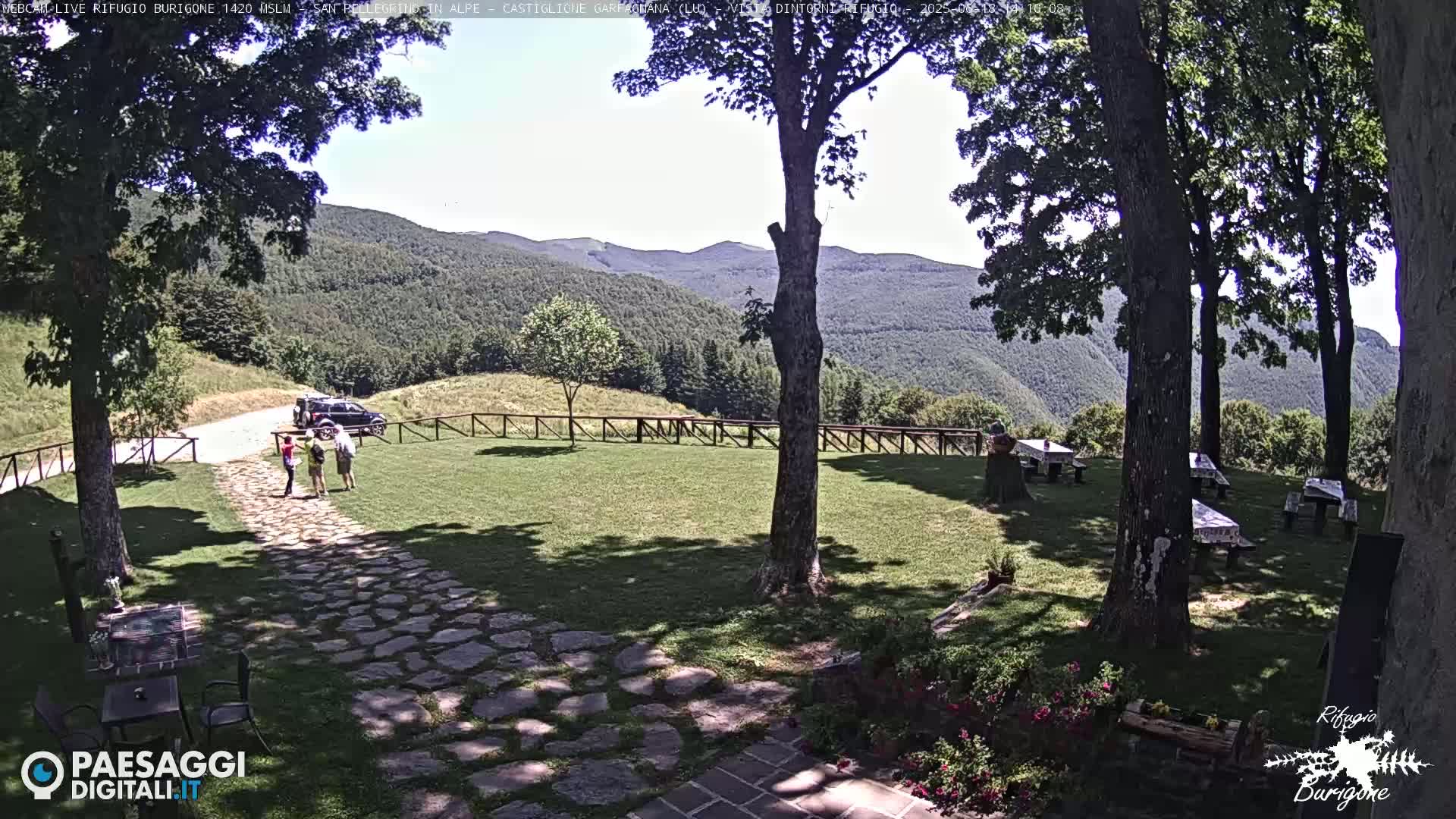 A stone path leads to a grassy area with picnic tables under trees, overlooking a mountainous landscape on a sunny day.
