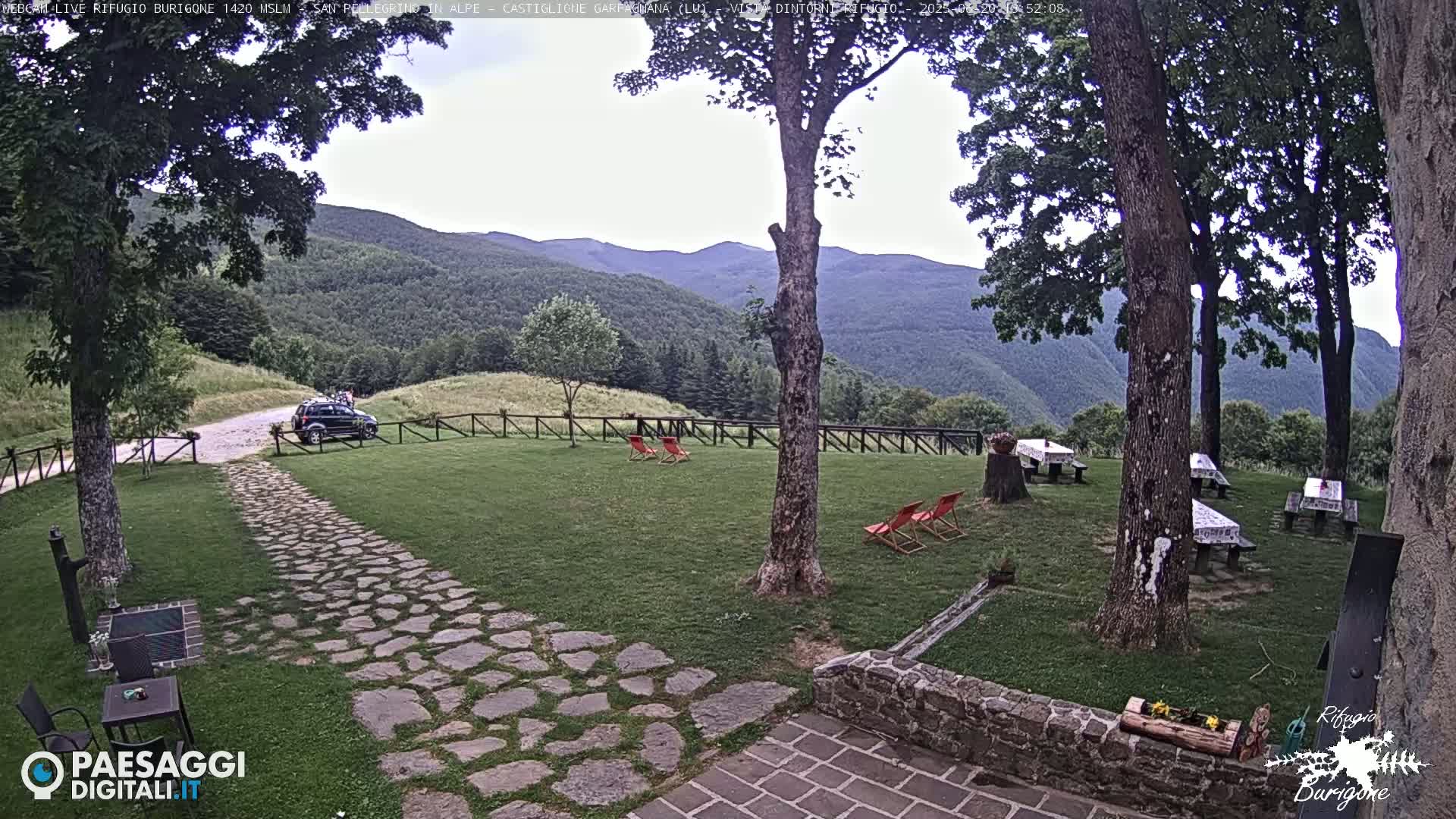 A stone path leads to a grassy area with several picnic tables and lounge chairs, all set against a backdrop of verdant hills under a mostly cloudy sky.