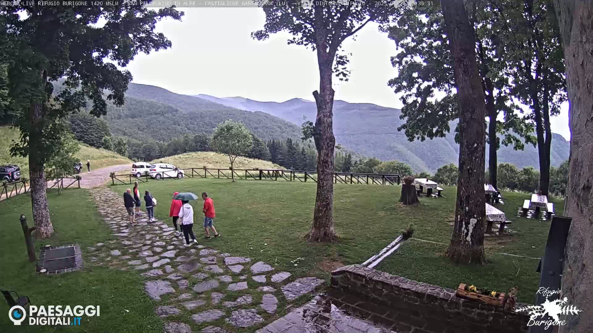 A stone path leads to several people walking under overcast skies toward picnic tables in a grassy area, with mountains in the background.