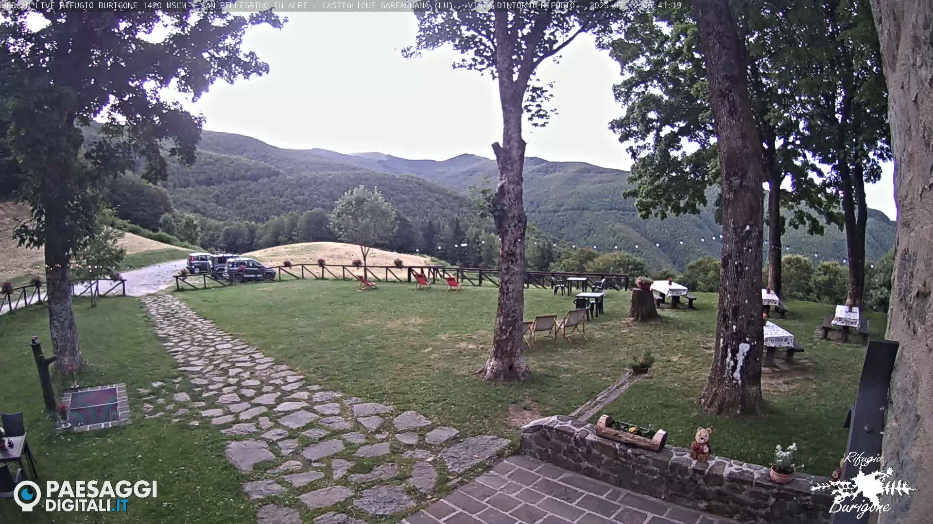 A stone path leads to picnic tables situated on a grassy area, overlooked by trees and hills under a sunny sky.