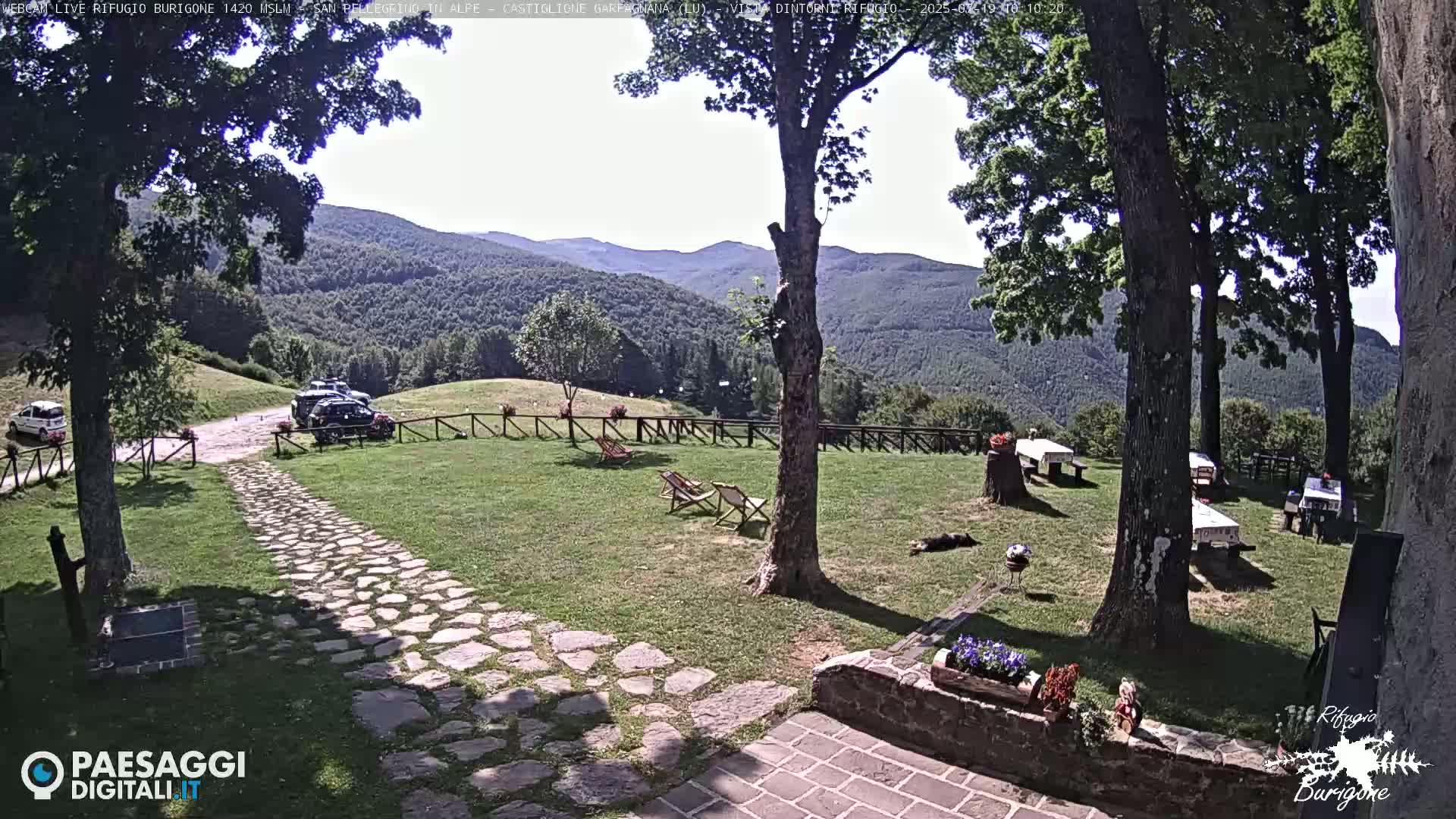 A stone path leads to a grassy area with several picnic tables and lounge chairs, overlooking a mountain range under a sunny sky.