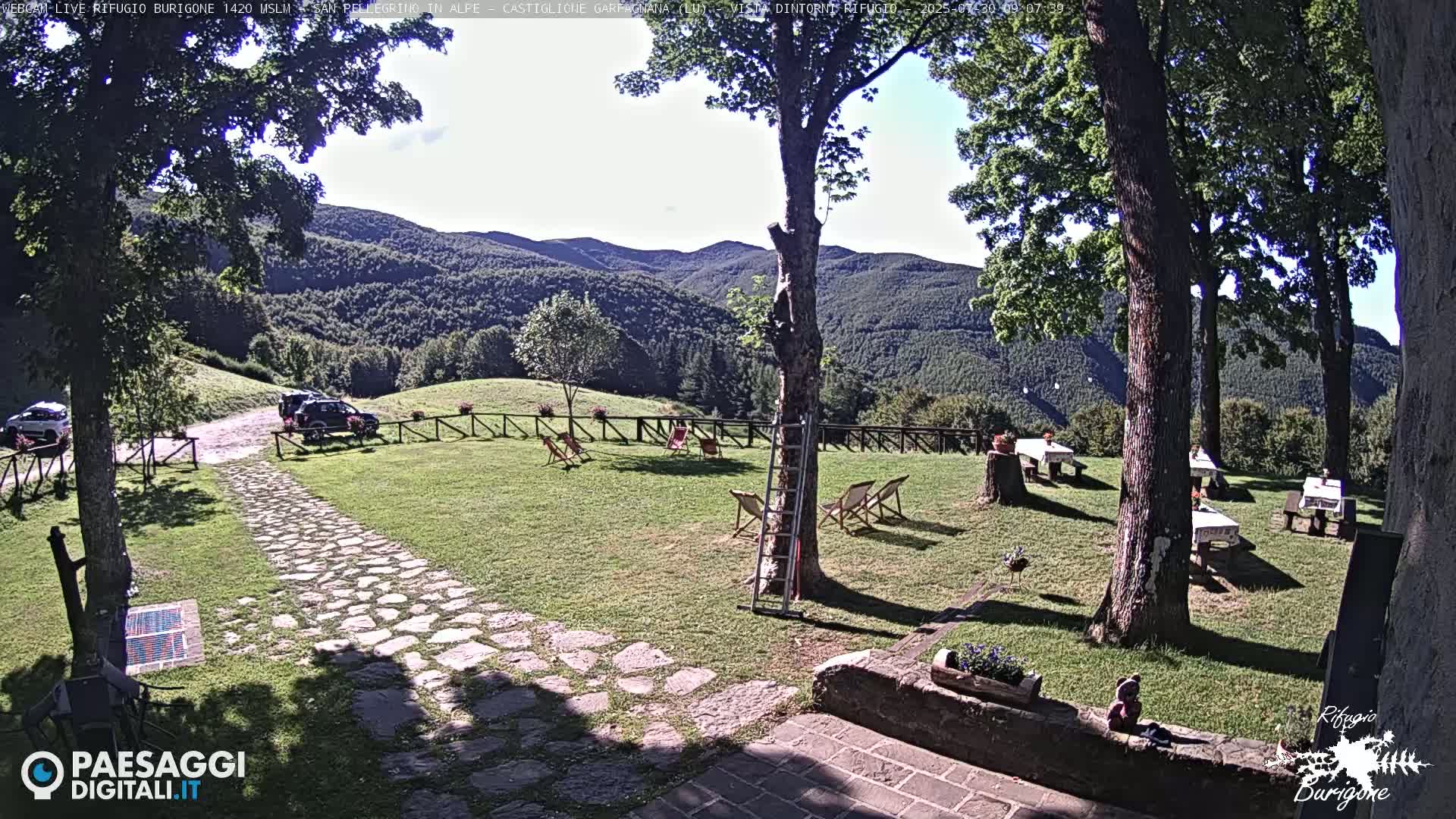 A stone path leads to a grassy area with several picnic tables, chairs, and a ladder, all situated in front of a mountainous landscape under a sunny sky.