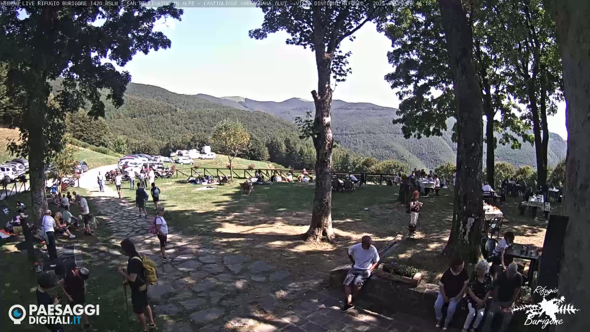 A stone path leads to a grassy area with several picnic tables, chairs, and a ladder, all situated in front of a mountainous landscape under a sunny sky.