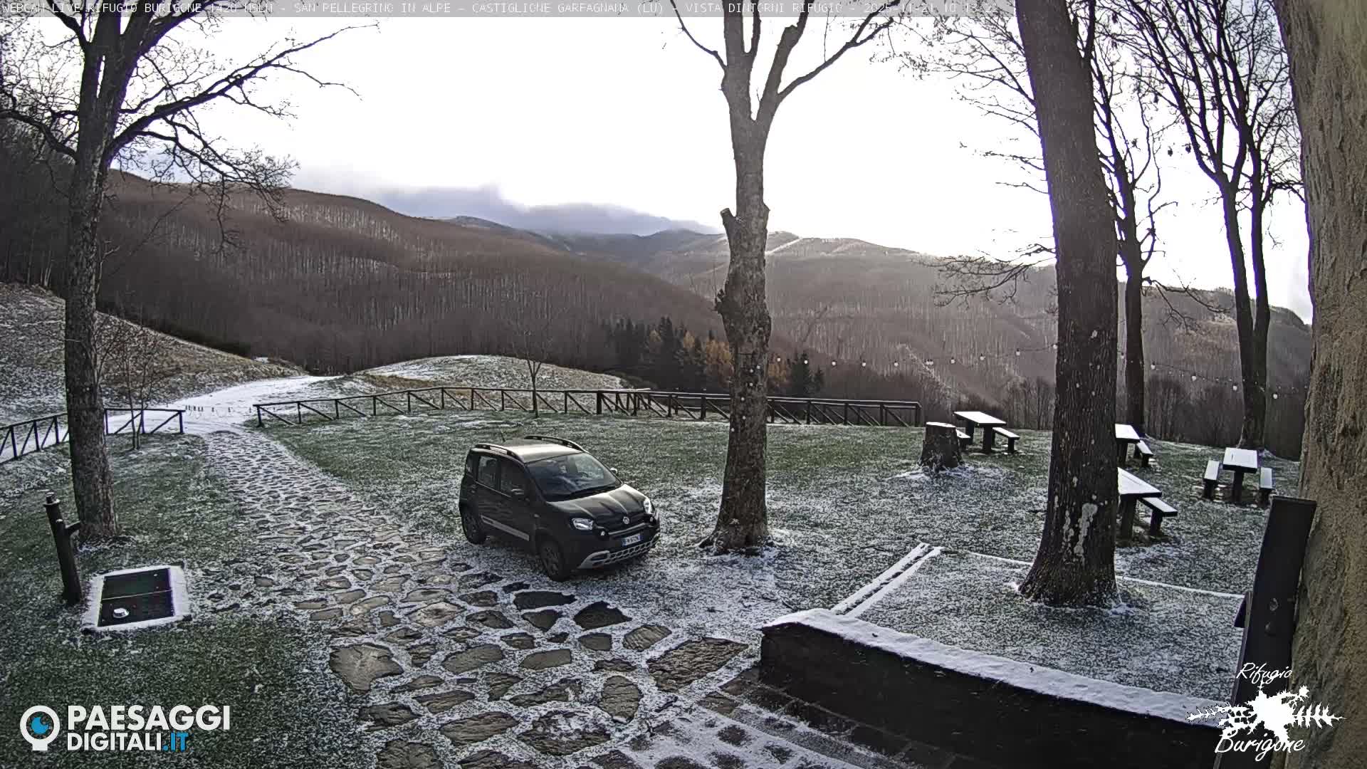 A light dusting of fresh snow covers the green grass and stone path where a dark car is parked, surrounded by bare trees and wooden picnic tables, with distant forested mountains visible under an overcast sky.