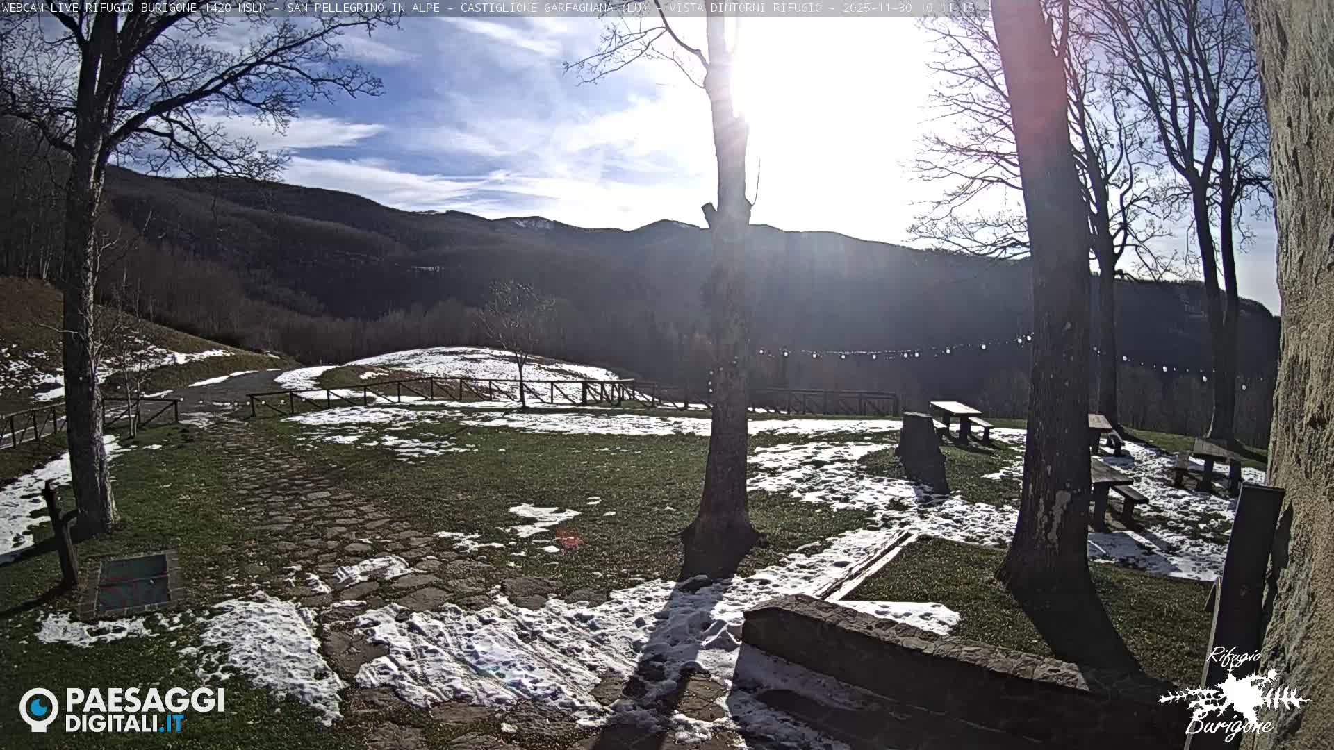 A sunny winter day illuminates a snowy, grassy clearing with bare trees and picnic tables, set against a backdrop of distant hills under a partly cloudy sky.