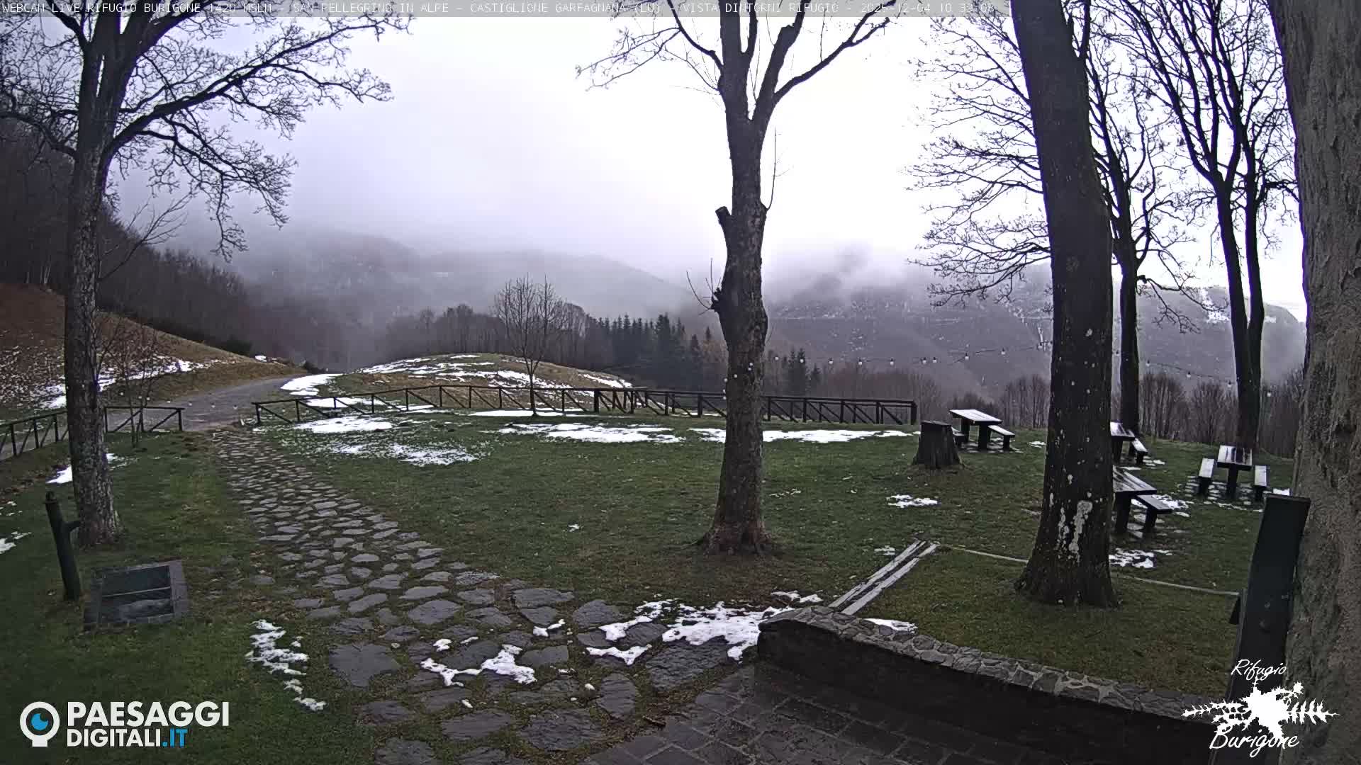 A foggy and overcast outdoor scene depicts a grassy area with bare trees, patches of snow, a cobblestone path, and several picnic tables, all set against a backdrop of mist-shrouded mountains.