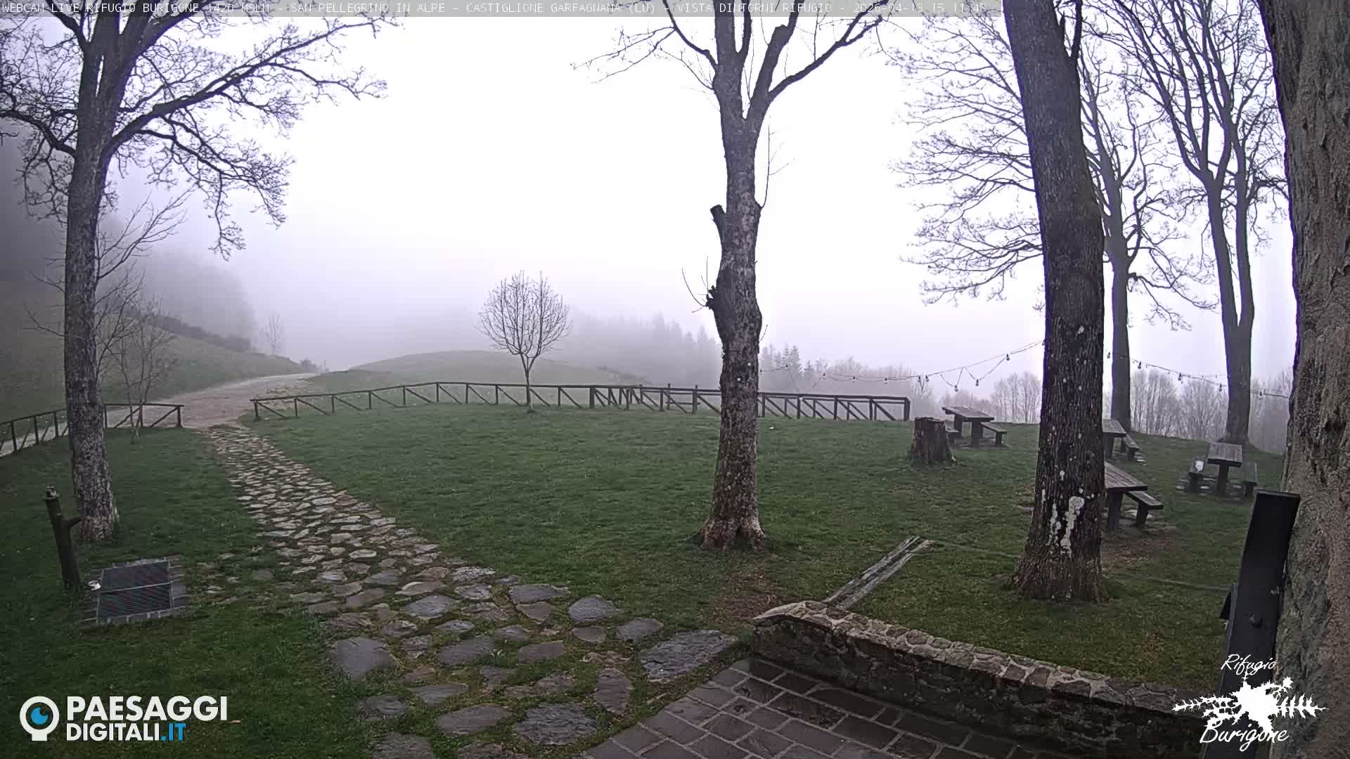 A foggy and overcast outdoor scene depicts a grassy area with bare trees, patches of snow, a cobblestone path, and several picnic tables, all set against a backdrop of mist-shrouded mountains.