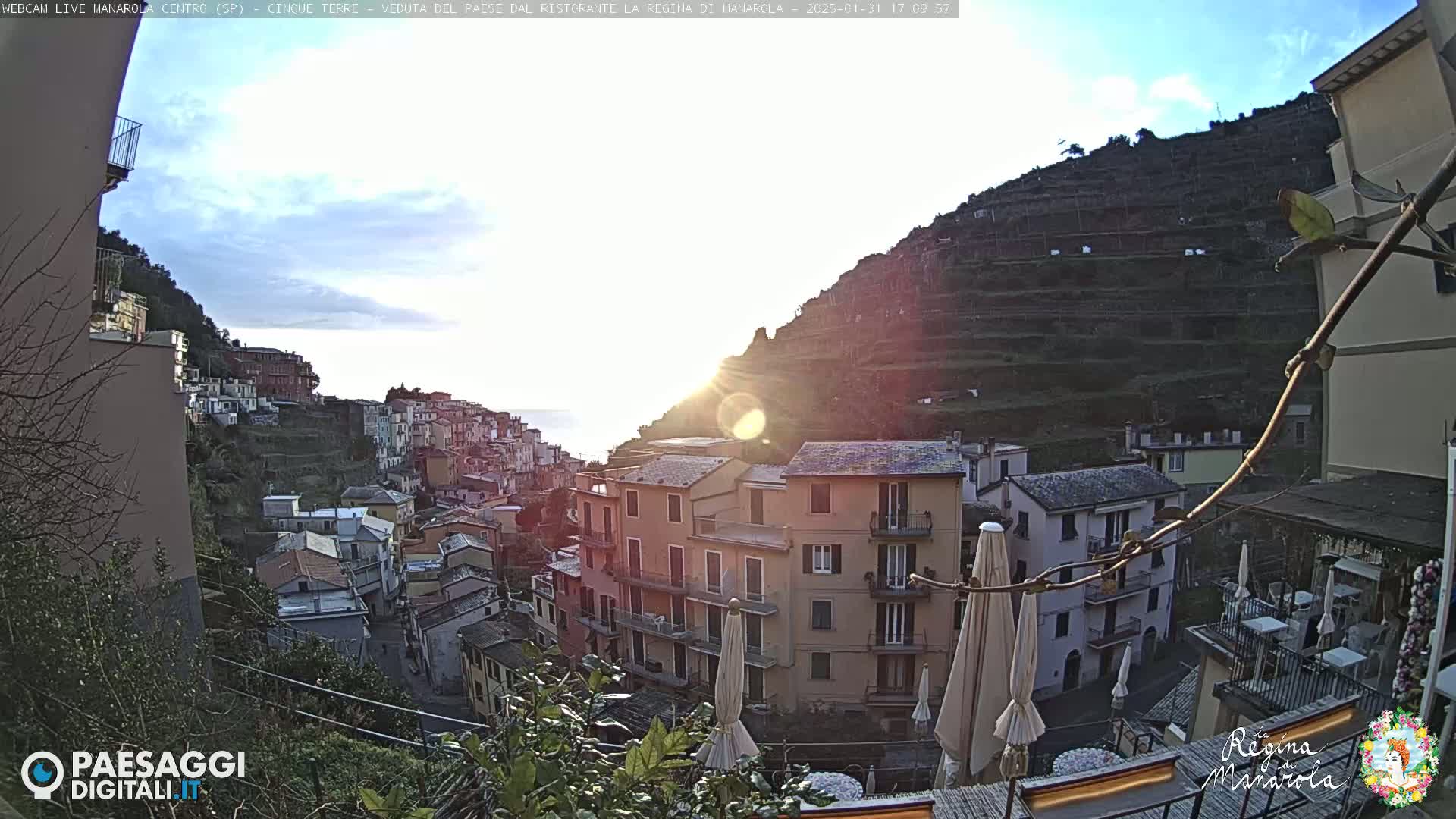 A sunlit view of a coastal town built into a hillside, with terraced fields rising behind it, under a mostly clear sky.