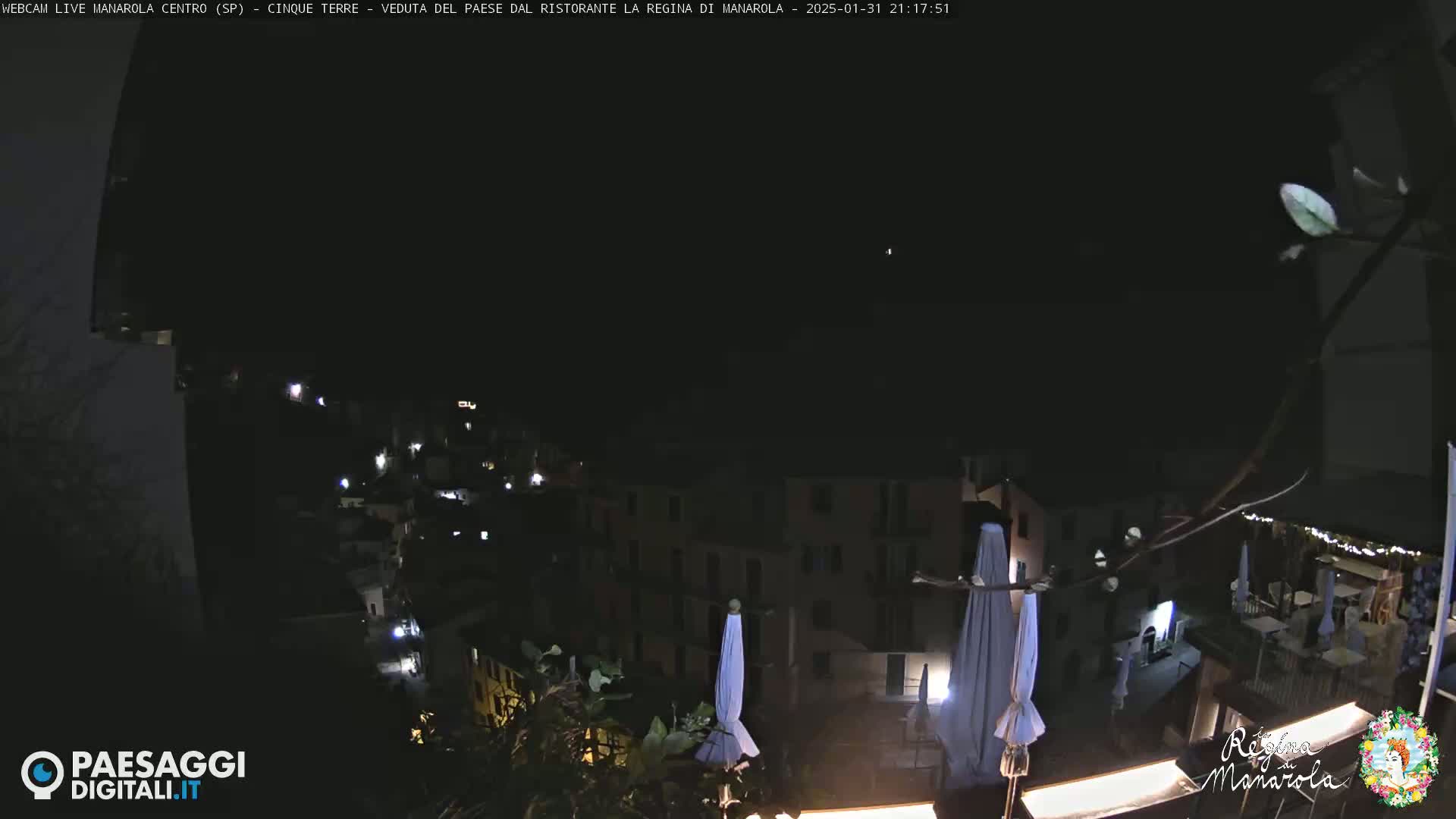 A nighttime view of a village nestled on a hillside, with several lights illuminating buildings and closed umbrellas visible in the foreground under a clear, dark sky.