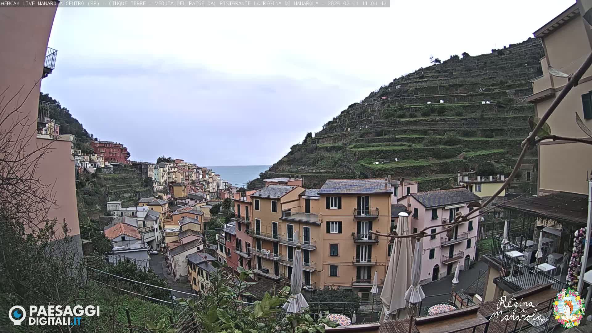 A coastal village of colorful buildings clings to a hillside overlooking a calm sea under an overcast sky.