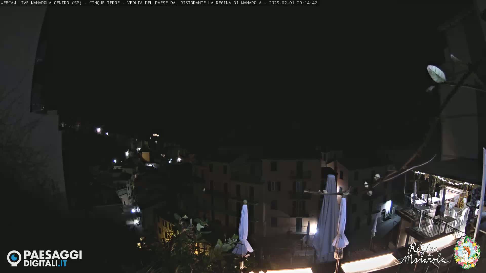 A nighttime view of a town built on a hillside, with several buildings illuminated and closed white umbrellas visible in the foreground.