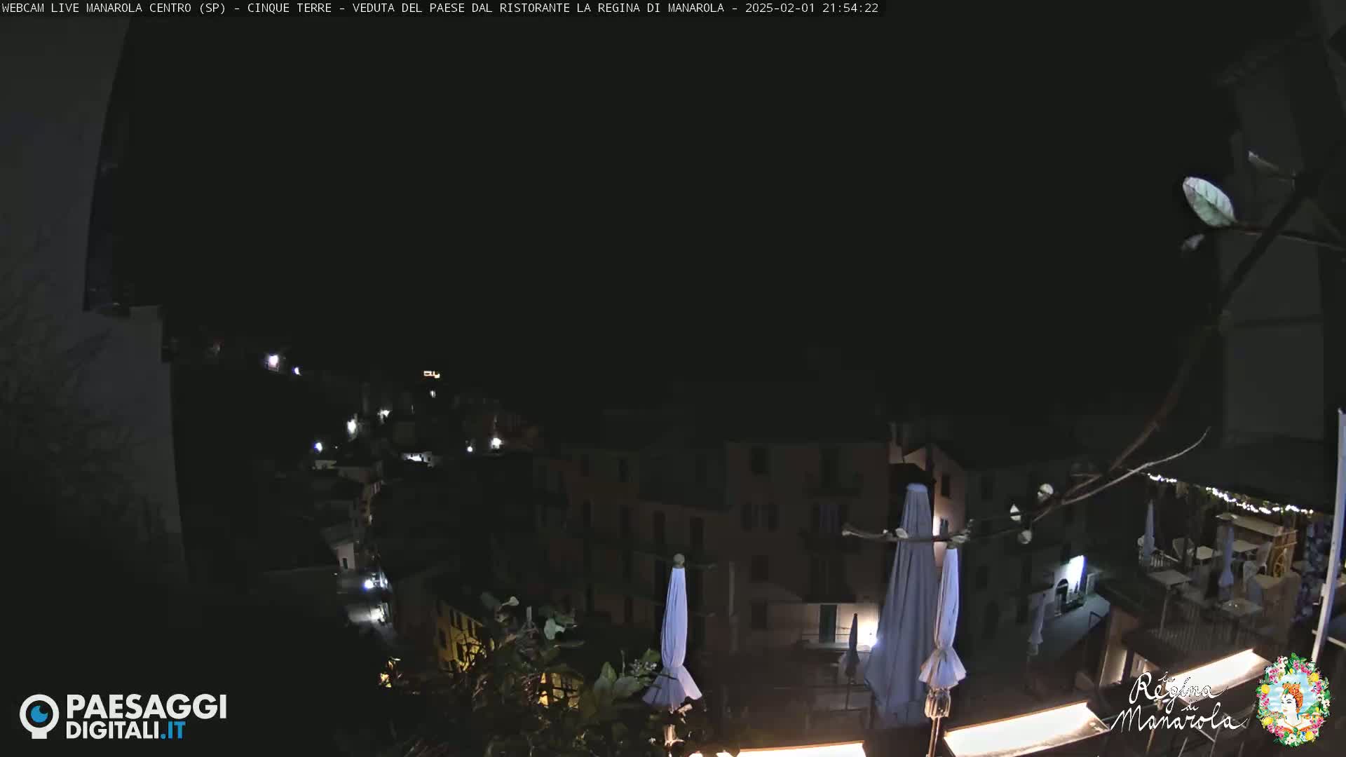 A nighttime view of a village, illuminated by scattered lights, with several closed outdoor umbrellas visible in the foreground.