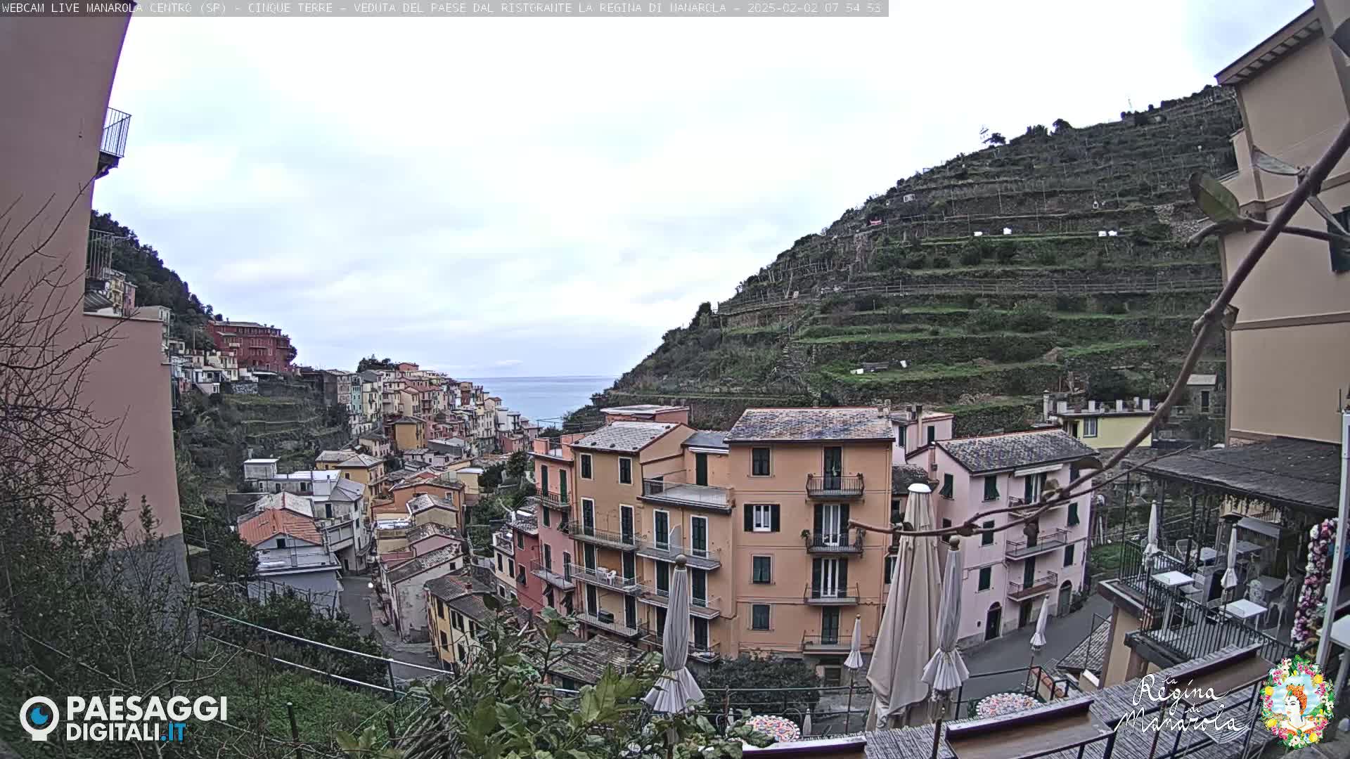 A coastal village of colorful buildings clings to a hillside overlooking a calm sea under an overcast sky.