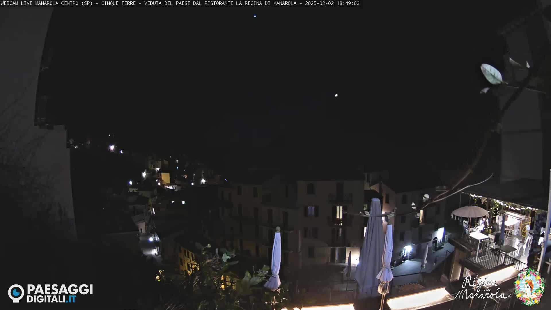 A nighttime, high-angle view of a village nestled on a hillside, with some lights visible and several closed umbrellas on a patio.